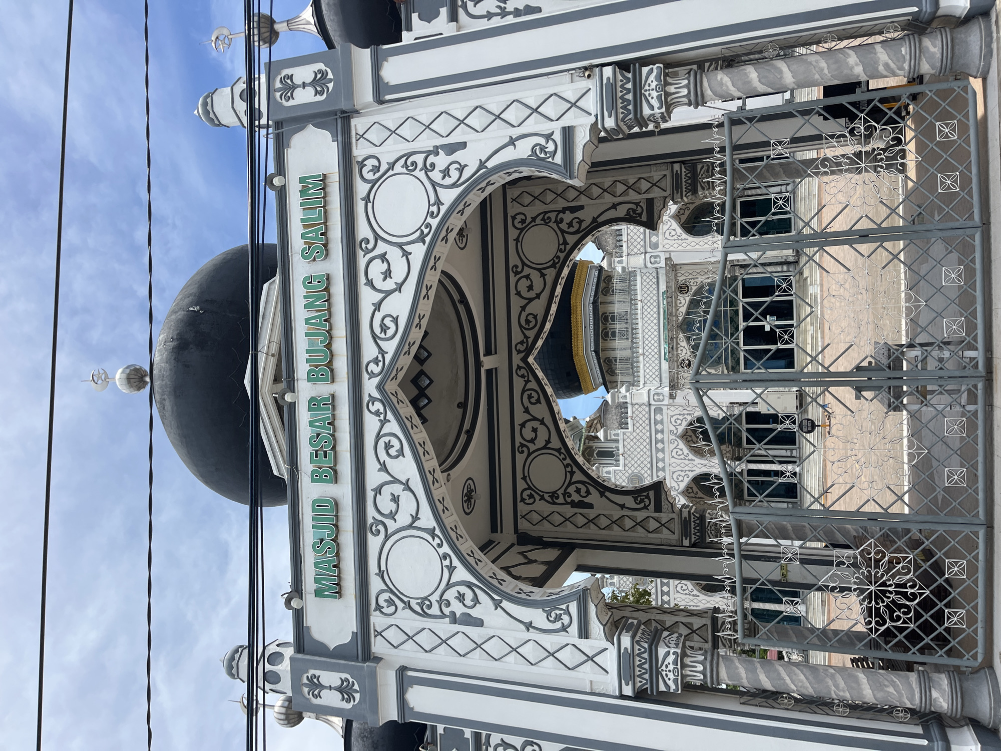 The image shows the Masjid Besar Bujang Salim mosque in Keudekruenggeukueh, Indonesia. The view is framed by a decorative arched entrance with Islamic-style patterns. The mosque's large black dome is visible above the entrance. The entrance is also gated, with a metal fence obstructing the view inside. Powerlines are seen cutting across the upper left of the image against the blue sky.