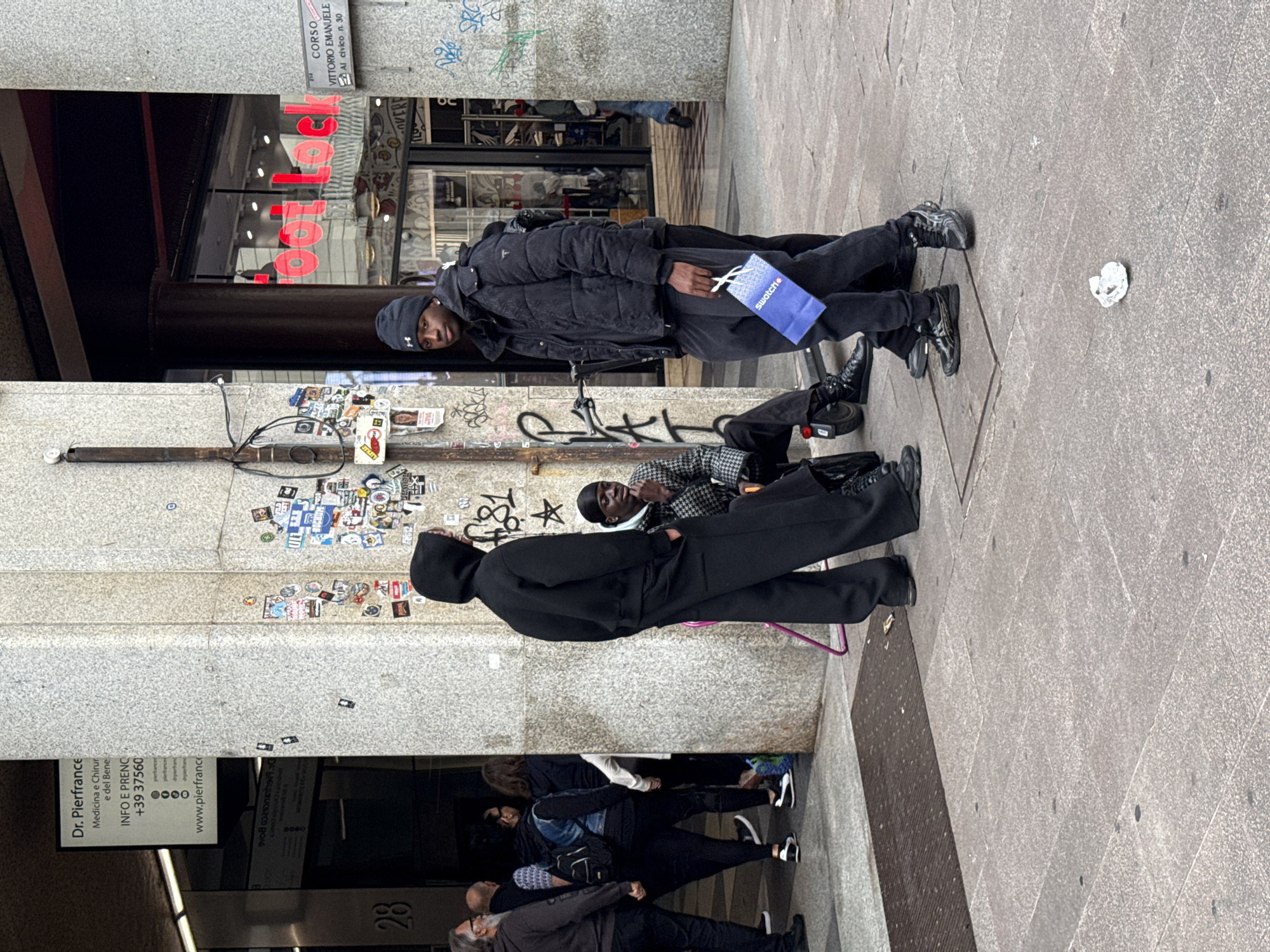 The scene shows a group of people on a street in Milan, Italy. Three individuals, one with a Swatch bag, are situated near a wall covered in stickers and graffiti. Others are walking past an entrance with a sign for "Dr. Pierfrance" and the address "Corso Vittorio Emanuele, Al civico n. 30," placing the location in the city center. One person is sitting, while another stands nearby. There is a discarded piece of paper on the pavement. The scene is urban, typical of a busy city street.
