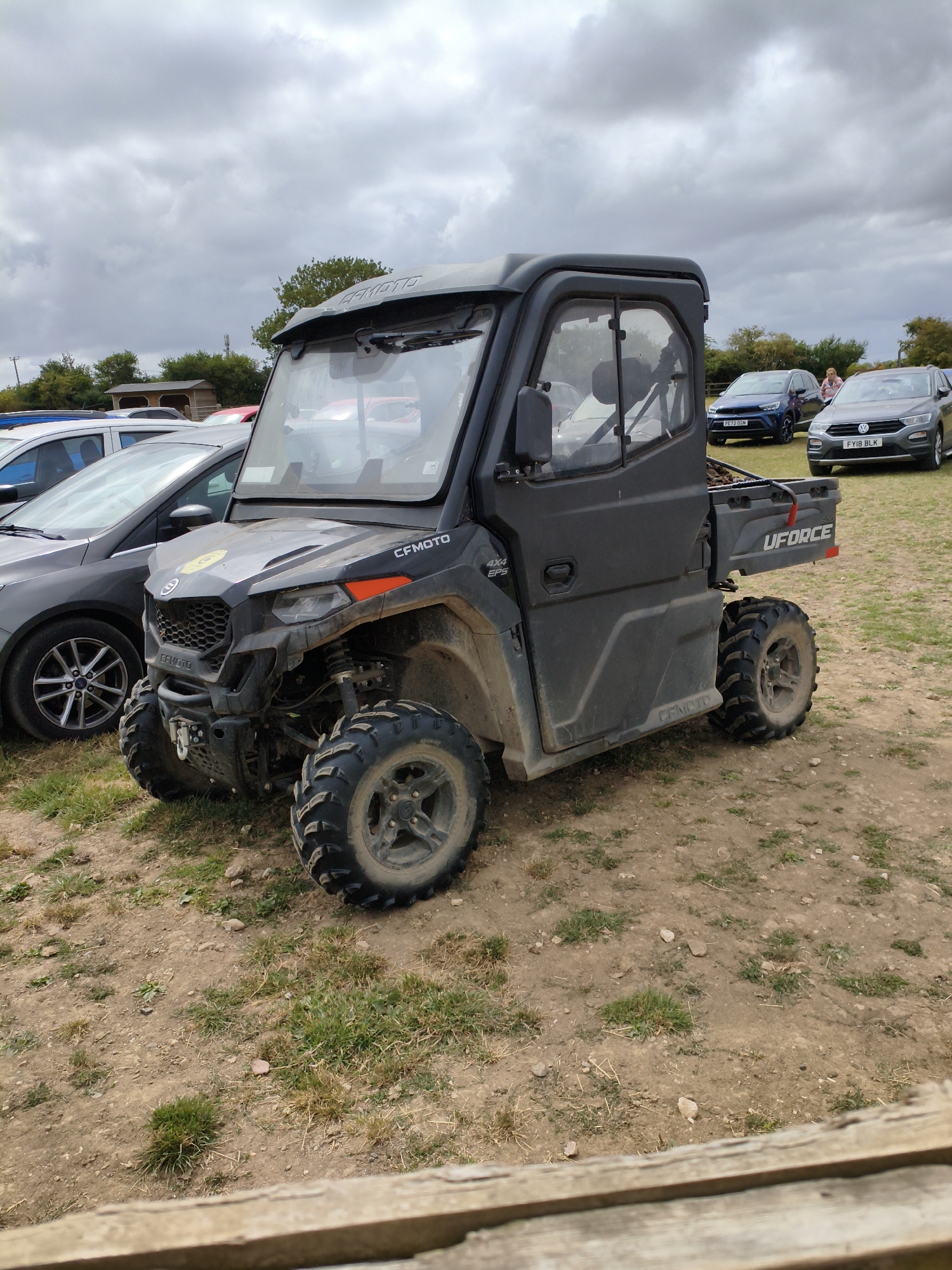 A black CFMOTO UFORCE utility vehicle is parked on a patch of dry, grassy ground under a cloudy sky. The vehicle has a cab with a windshield, doors, and a roof. In the background, several cars are parked, including a dark gray Volkswagen T-Roc with the license plate FY18 BLK. The scene appears to be a parking area, possibly at an outdoor event or recreational site, given the presence of the utility vehicle and the casual arrangement of cars. The environment is rural, with trees and open ground visible.