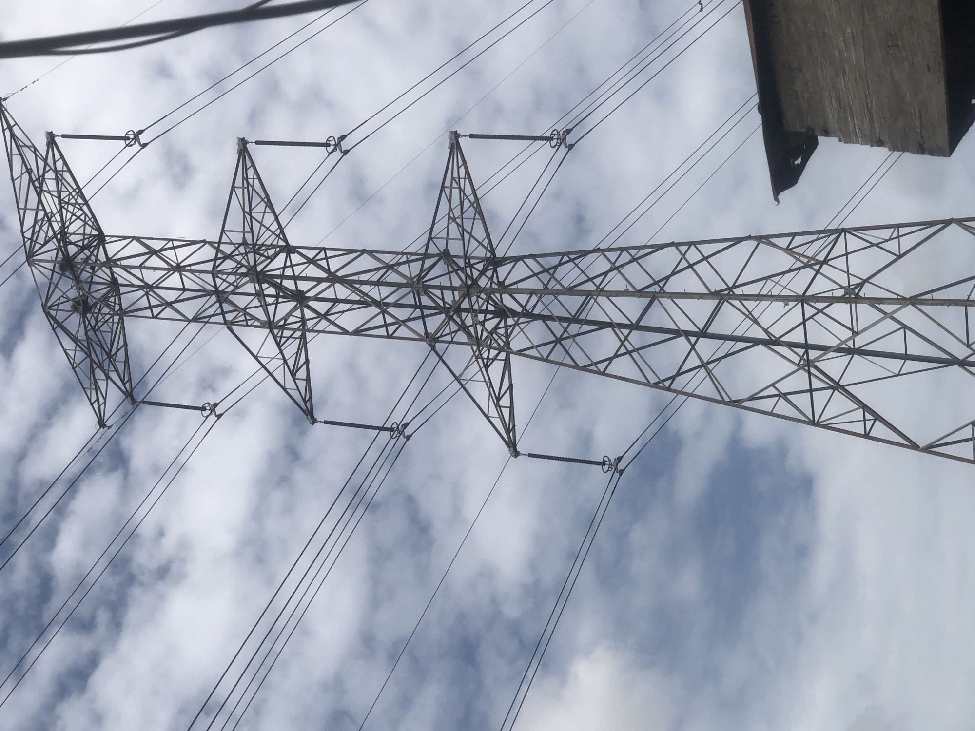 A large electricity transmission tower, commonly known as a pylon, dominates the frame, viewed from below against a bright, partly cloudy sky. The pylon is constructed from a robust lattice framework of metal beams, forming intricate geometric patterns. Multiple high-voltage power lines extend across the upper section, suspended from the pylon's horizontal arms by ceramic or composite insulators, which appear as stacked discs.
The sky in the background exhibits a mix of white and light grey clouds, indicative of daytime and fair weather. In the top right corner, a dark, solid, and partially visible structure, possibly a concrete bridge or an elevated section of a building, juts into the scene. The photograph captures a static moment of infrastructure. There are no people or specific activities depicted, and no visible text can be identified on any of the objects. The scene represents a typical electrical grid component in Ado, Nigeria.