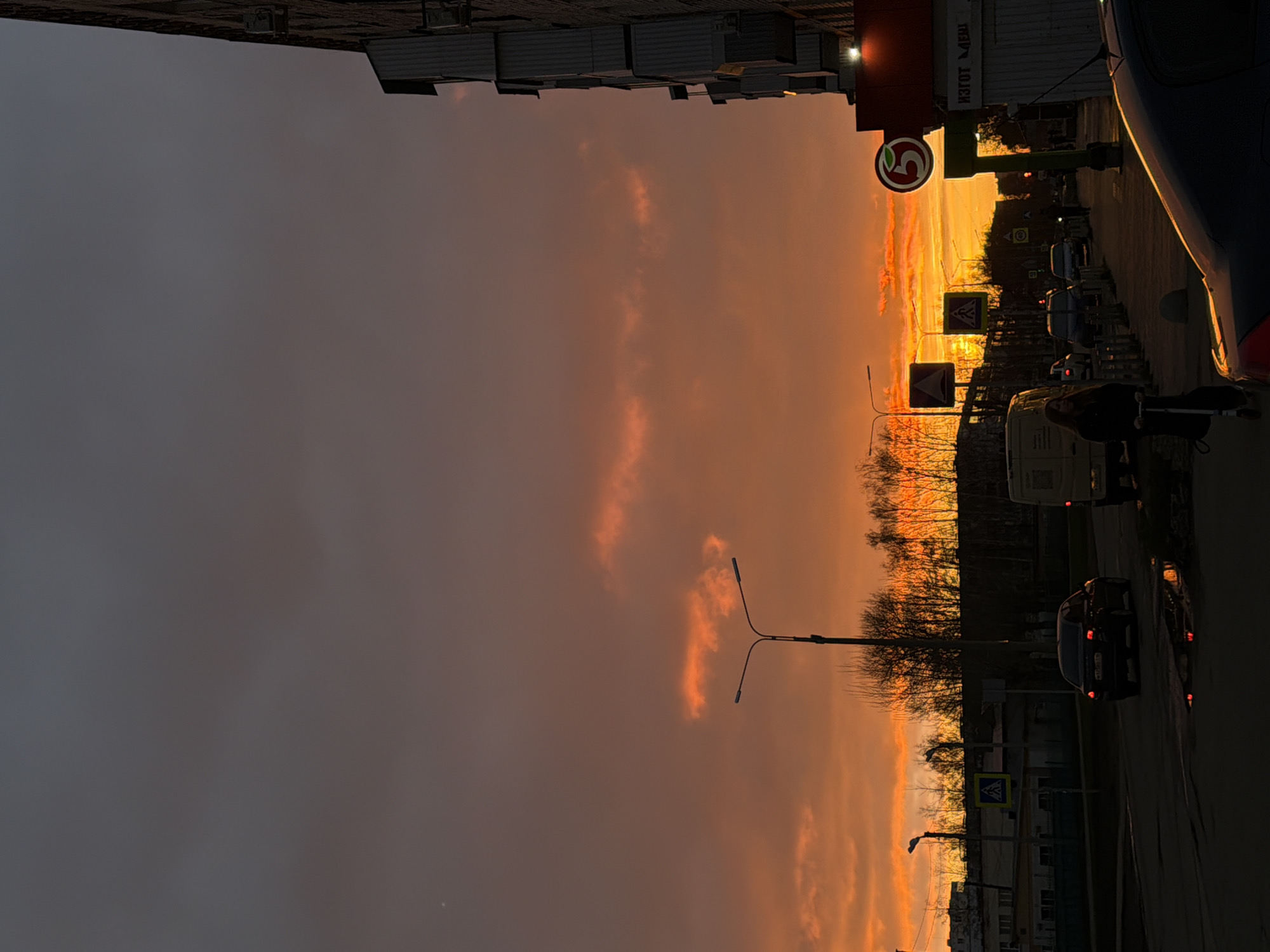 This media file captures an urban street scene at sunset in Naberezhnye Chelny, Russia, bathed in warm, dramatic light. The sky, especially prominent in the upper and left parts of the image, displays vivid shades of orange, red, and yellow, indicating the setting sun. Some darker clouds are also visible, contrasting with the brightly illuminated portions of the sky.
Along the right side of the frame, an asphalt road features various vehicles. A dark car is partially visible in the foreground, and other cars and a white van are further down the street. A person is seen riding a bicycle or scooter on the right side of the road. Several lampposts are silhouetted against the bright sky, extending over the street. Traffic signs, including a blue square pedestrian crossing sign and a triangular warning sign, are present.
On the right, a building is adorned with a distinctive red and green sign featuring a white number "5" logo, strongly suggesting a Pyaterochka supermarket. Another building in the background has faintly visible Russian text, possibly "ИЗГОТ МВИ" or similar. Bare-branched trees, likely indicating an autumn or winter season, are silhouetted against the colorful horizon. In the distance, dark urban buildings complete the scene. The overall impression is a bustling city street transitioning from day to night, highlighted by the striking colors of the sunset.