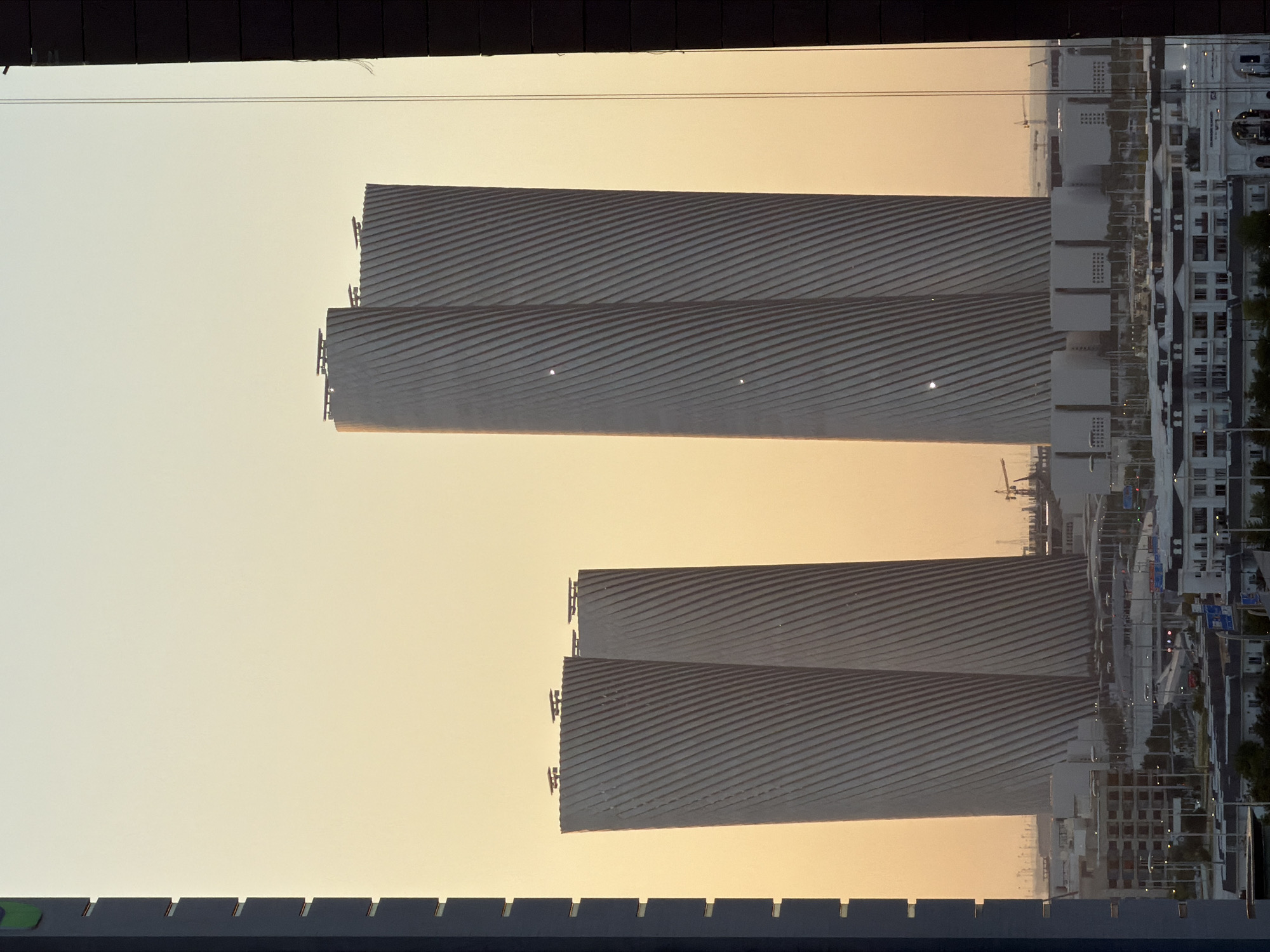 A dusk or dawn view of the twisted towers of the Al Bayt Stadium in Al Khor, Qatar, is framed by the dark vertical structures of other buildings on the left and right. The sky is a pale gradient of yellow and orange. The iconic stadium, designed to resemble a nomadic tent, is captured from a distance, its distinctive white, curved surfaces featuring diagonal lines. Smaller buildings and infrastructure, including streetlights and what appear to be lower-rise structures, are visible in the foreground at the base of the stadium. Construction cranes are also present in the background, indicating ongoing development in the area. The overall scene conveys an architectural and urbanistic landscape under a soft, atmospheric sky.