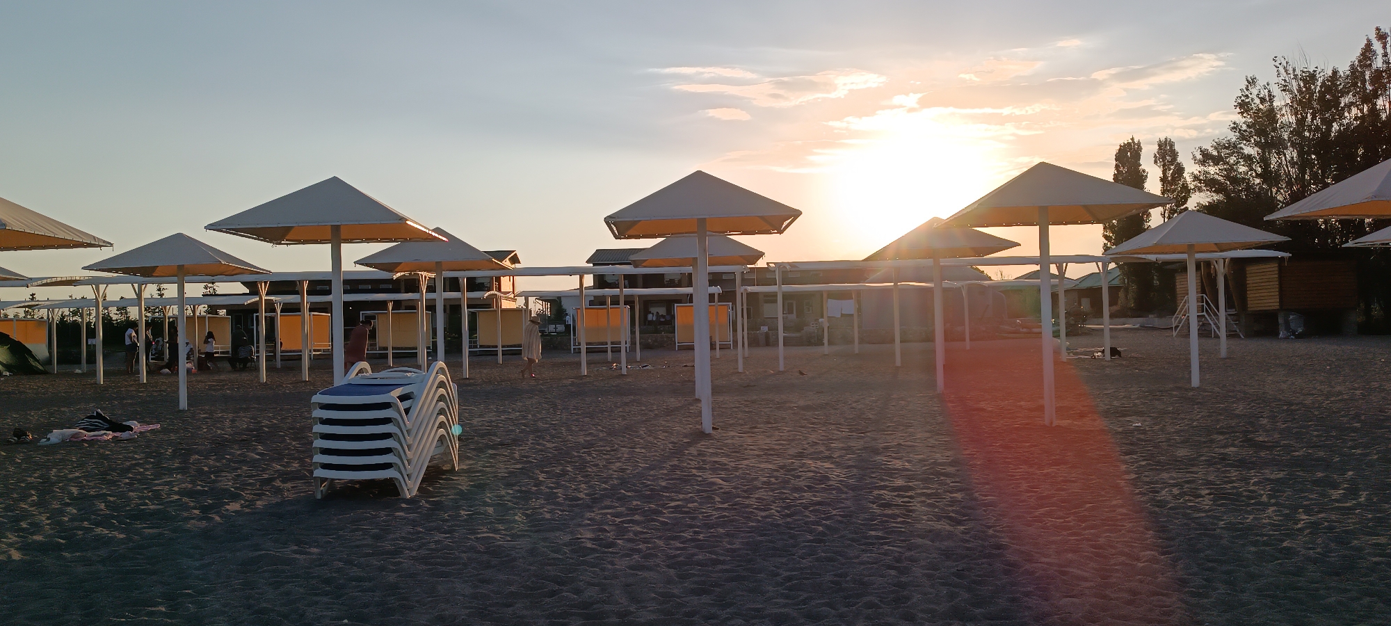 An outdoor, low-angle, wide shot captures a sandy beach area in Koshkol', Kyrgyzstan, during sunset. The sky is a clear gradient of pale blue and orange, with the sun partially obscured by clouds and casting a warm glow. Several white beach umbrellas with yellow undersides are erected across the sand, casting shadows. In the foreground, a stack of white and blue lounge chairs sits on the left side. To the left of the chairs, two people are lying on blankets on the sand. In the middle ground, a few individuals are gathered around and walking between the umbrellas. In the background, there are buildings with yellow awnings, trees, and structures that appear to be cabanas or small shops. The overall atmosphere is tranquil and depicts a typical beach scene in the late afternoon.