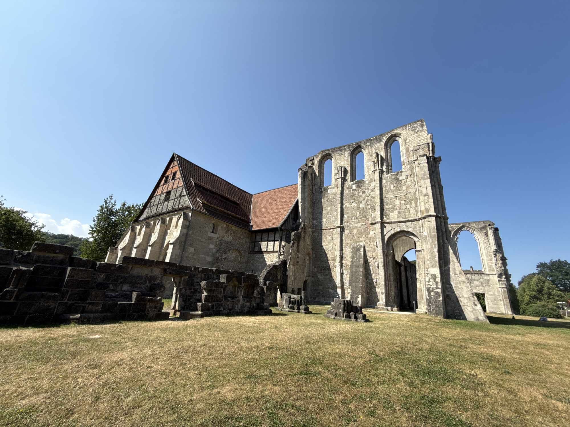 The image depicts the remains of the Walkenried Abbey in Walkenried, Germany, under a clear blue sky. The foreground is dominated by dry, brown grass, with several low stone walls and structures scattered across the field. In the mid-ground, a partially preserved stone building stands, featuring arched window openings and weathered stone architecture. To the left of this structure, a more intact building with a reddish-brown tiled roof and timber framing is visible. The overall scene conveys a sense of historical ruin and preservation within a natural, outdoor environment. There are no people visible in the image.