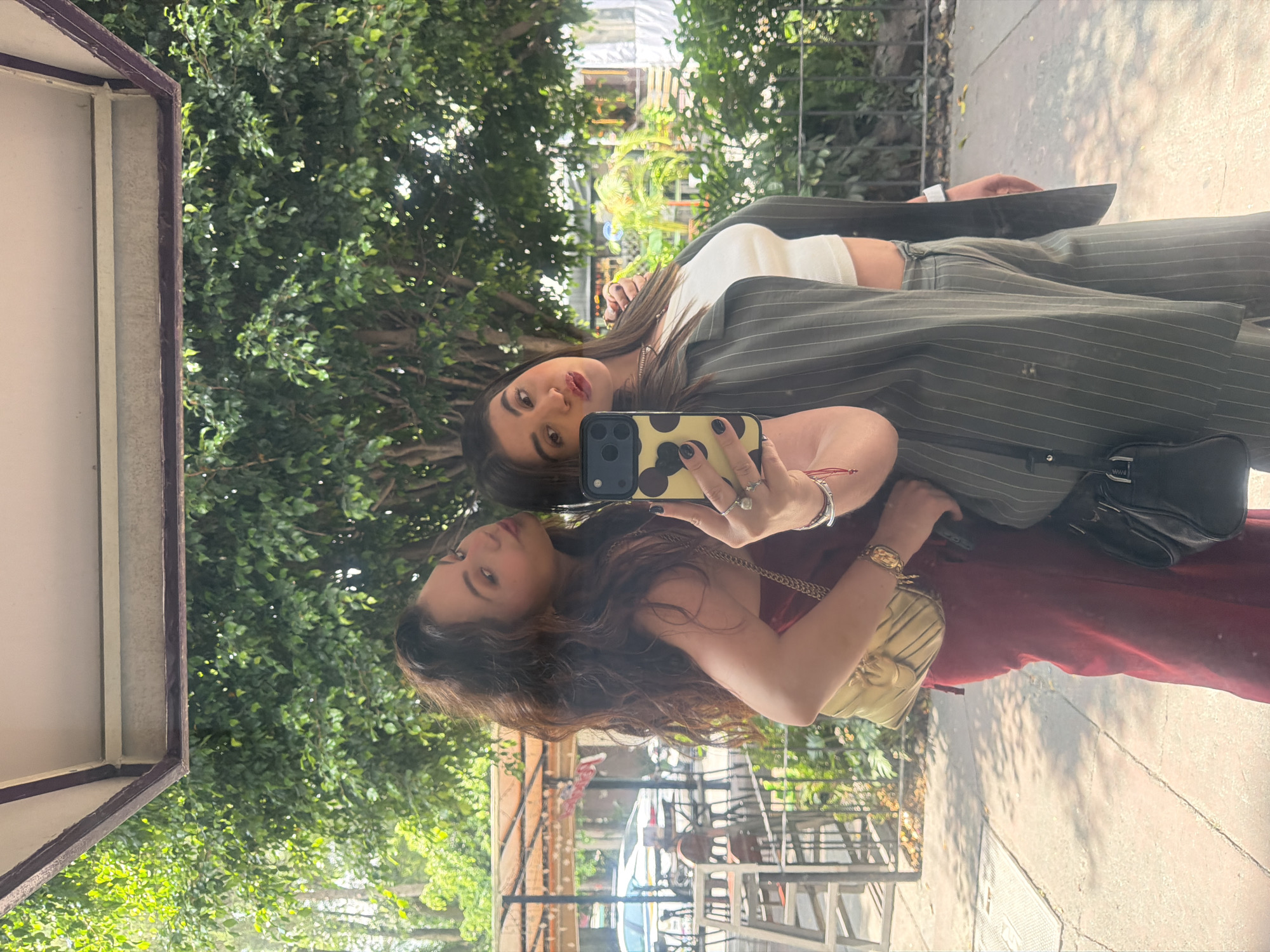 A photograph taken in Mexico City shows a young woman's reflection in a large window. The woman appears to be taking a selfie with a smartphone with a yellow and black patterned case. She is wearing a striped blazer over a white top and matching pants. She has dark hair and red lipstick. In the reflection, a second woman with dark, wavy hair wears a red dress and a gold handbag. The background of the photo features lush green trees and a street scene with cars and buildings. The location is identifiable by the typical architecture and vegetation commonly found in Mexico City.