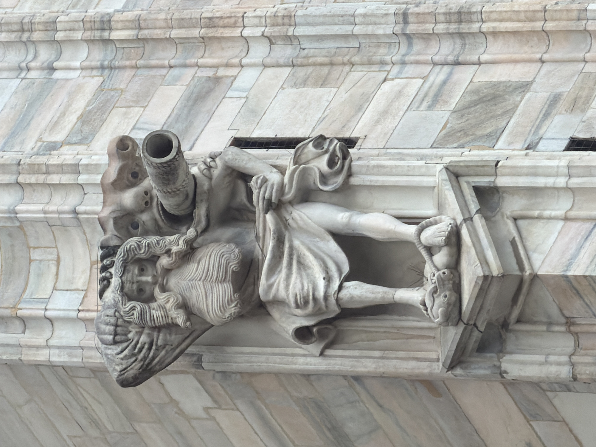 A detailed marble statue, likely a gargoyle or grotesque, is prominently featured, affixed to the facade of a large building. The statue depicts a male figure with a contorted, grotesque face, characterized by deeply set eyes, a prominent nose, and an open mouth. Long, wavy hair or a beard flows around its head and shoulders. A large, cylindrical spout protrudes from the figure's head or neck area, suggesting its function to channel water.
The figure is carved in a dynamic, somewhat reclining pose, with drapery covering its lower body. Its hands are visible near its lower extremities, appearing to hold or interact with a small animal's head, which resembles that of a rabbit. The carving exhibits intricate detail in the hair, drapery, and the figure's overall form.
The background consists of a finely crafted marble wall, displaying horizontal and diagonally arranged blocks with natural light grey and white veining. Some areas show signs of age and weathering. The bright, even lighting suggests the photo was taken on a clear day. The architectural style and material are consistent with significant historical buildings in Milan, Italy, such as the Duomo di Milano, known for its numerous statues and use of Candoglia marble. No text is visible in the image.
