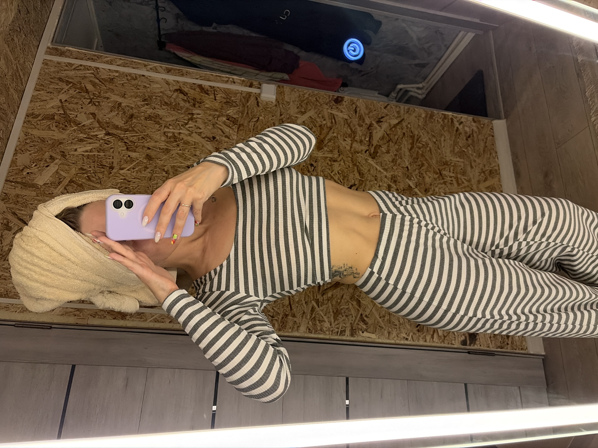An adult individual stands in front of a mirror, taking a selfie within an interior space in the city of Chaikovsky, Russia. Their face is largely obscured by a light purple smartphone held in their right hand and their left hand covering the upper face/forehead. A light beige terry cloth towel is wrapped around their head. The individual wears a two-piece horizontally striped ribbed knit outfit, consisting of a long-sleeved crop top and matching pants, exposing the midriff. Tattoos are visible on the left side of the torso and near the right collarbone. The nails on the right hand are painted white, with some featuring colorful abstract designs; a thin gold ring is on the right ring finger.
The mirror's reflection reveals the environment: to the right of the individual, a wall covered in Oriented Strand Board (OSB) is visible, featuring several small white rectangular fixtures, possibly outlets or switches. To the left, a light gray panel wall with dark horizontal lines is reflected. The floor is composed of light brown wooden planks, separated from the OSB wall by a white baseboard. In the far right of the reflection, a section of hanging garments and a circular object with a glowing blue power symbol are present. A vertical light strip or illuminated frame element is visible along the left edge of the mirror.