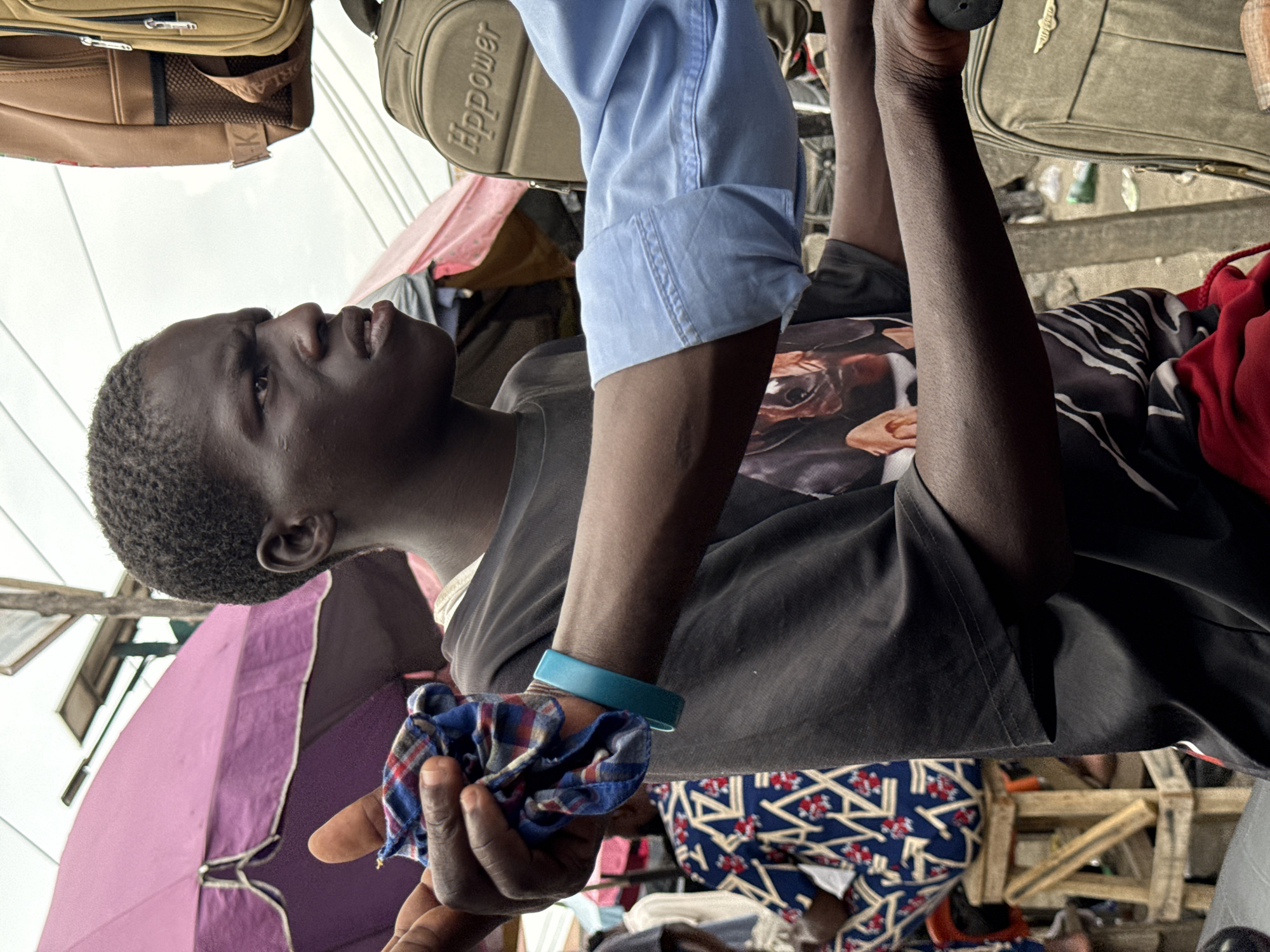 A dark-skinned young man stands in a bustling market in Lagos, Nigeria. He is wearing a black t-shirt with a printed design, a blue bracelet, and is holding a folded blue and red plaid cloth in his left hand. His face is visible in profile, and he seems to be looking upwards. A light blue shirt sleeve is visible near his arm.
The background reveals a chaotic marketplace setting. Several bags and suitcases are positioned above him, potentially hanging for sale or display. There's a purple umbrella providing shade, and various fabrics and materials are visible in the background, suggesting a vibrant commercial area. Other people are present, but they are partially obscured by the market goods and the angle of the photo. A makeshift wooden frame structure can be observed in the background.