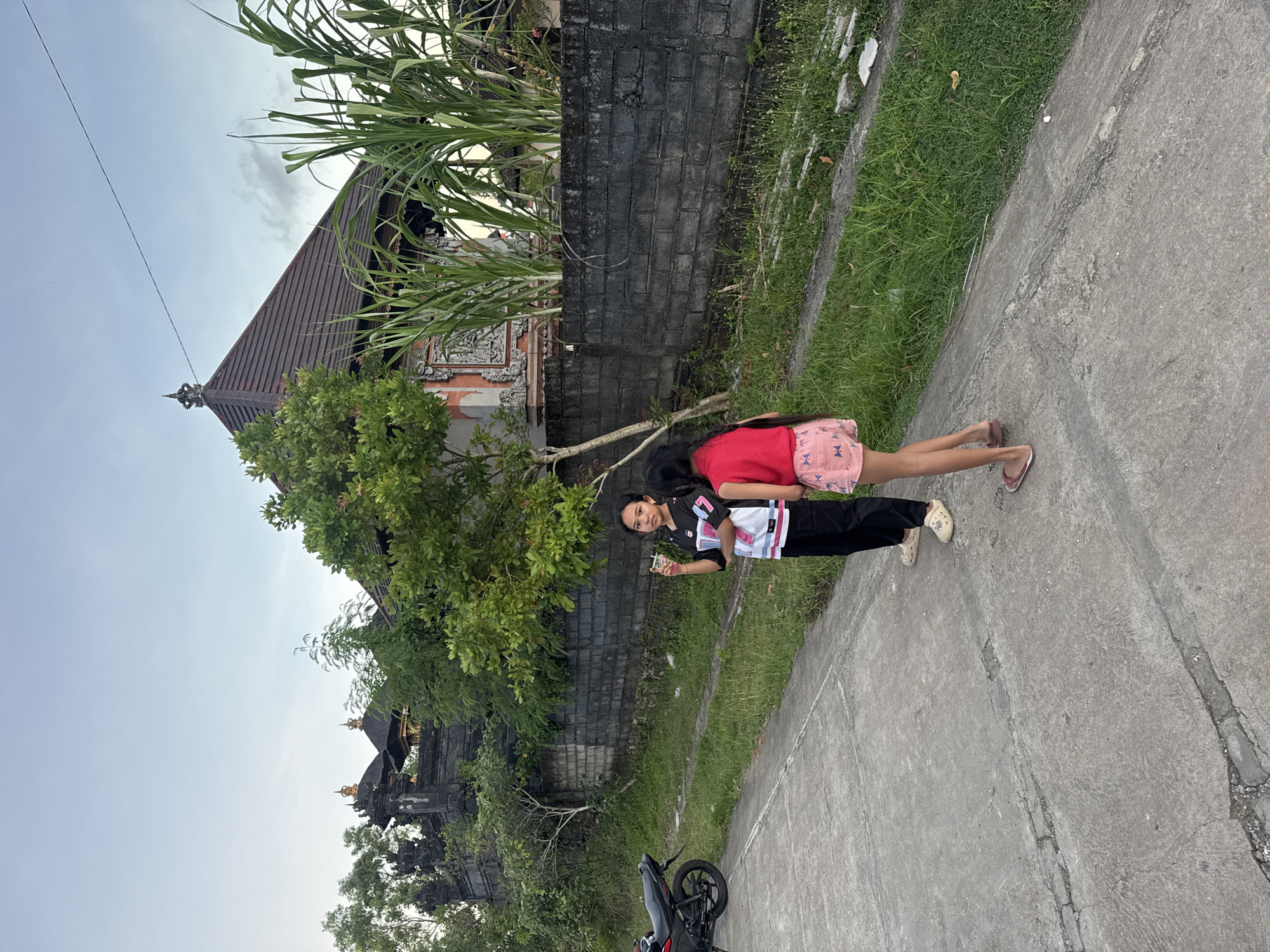 Two people, one wearing a black sports jersey and black pants and the other wearing a red t-shirt and pink shorts, stand on a concrete road adjacent to a brick wall. A motorcycle is parked on the road. Behind the wall is a temple-like building with tiered roofing. The location is Kuta Selatan, Indonesia.