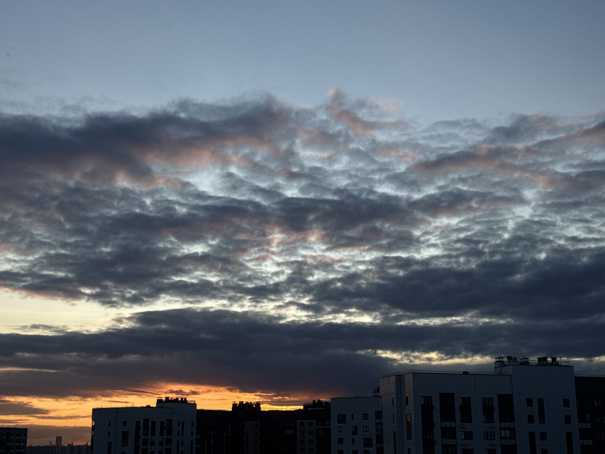 The image shows a cityscape at dusk or dawn, with a dramatic sky filled with dark, textured clouds tinged with pink and orange near the horizon. Silhouetted against this sky are the upper floors of several modern, multi-story buildings. These buildings are predominantly white or light-colored, with visible windows and rooftop structures such as ventilation units and antennae. The lighting suggests low natural light conditions, possibly indicating a time of day when artificial lights in the buildings might be beginning to turn on, though none are clearly illuminated in this shot. The overall scene depicts an urban environment under a brooding sky.