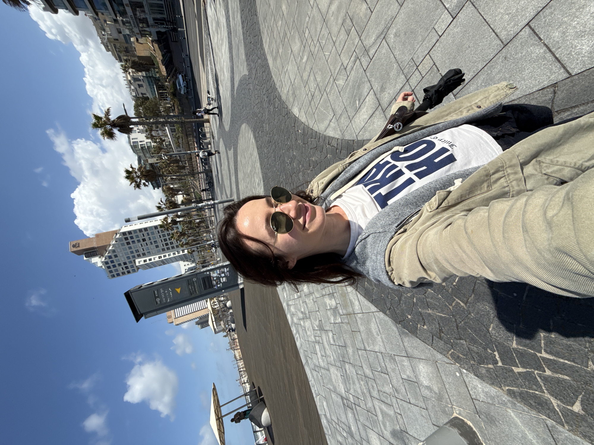 A medium-shot, canted-angle photograph captures a person wearing sunglasses, a white t-shirt with blue lettering, a gray hoodie, and a tan jacket. The individual is positioned on a paved area with a combination of gray rectangular pavers and curved patterns of smaller, dark-colored stones. The person's legs are visible and are draped with an umbrella. The backdrop is a cityscape with buildings, palm trees, and a clear blue sky with scattered clouds. A bus stop sign is noticeable in the background. The scene is set in the city of Tel-Aviv, Israel.