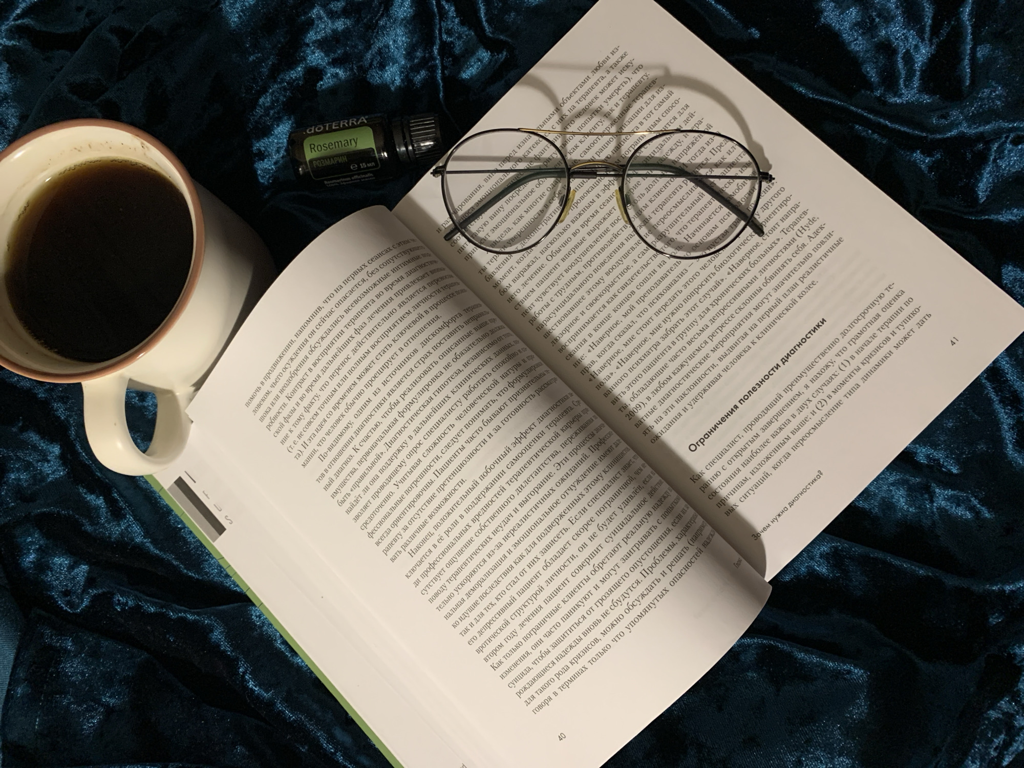 The image depicts a book lying open on a textured blue surface, possibly velvet. A pair of round-frame glasses rests on the right-hand page of the book, slightly askew. The pages appear to be filled with text in Cyrillic script. Next to the book, on the left side of the image, is a white ceramic mug filled with a dark liquid, likely coffee or tea. A small bottle of Rosemary essential oil with Cyrillic text is adjacent to the mug. The environment suggests an indoor setting with soft, ambient lighting, emphasizing the texture of the blue fabric and the warmth of the mug's contents.