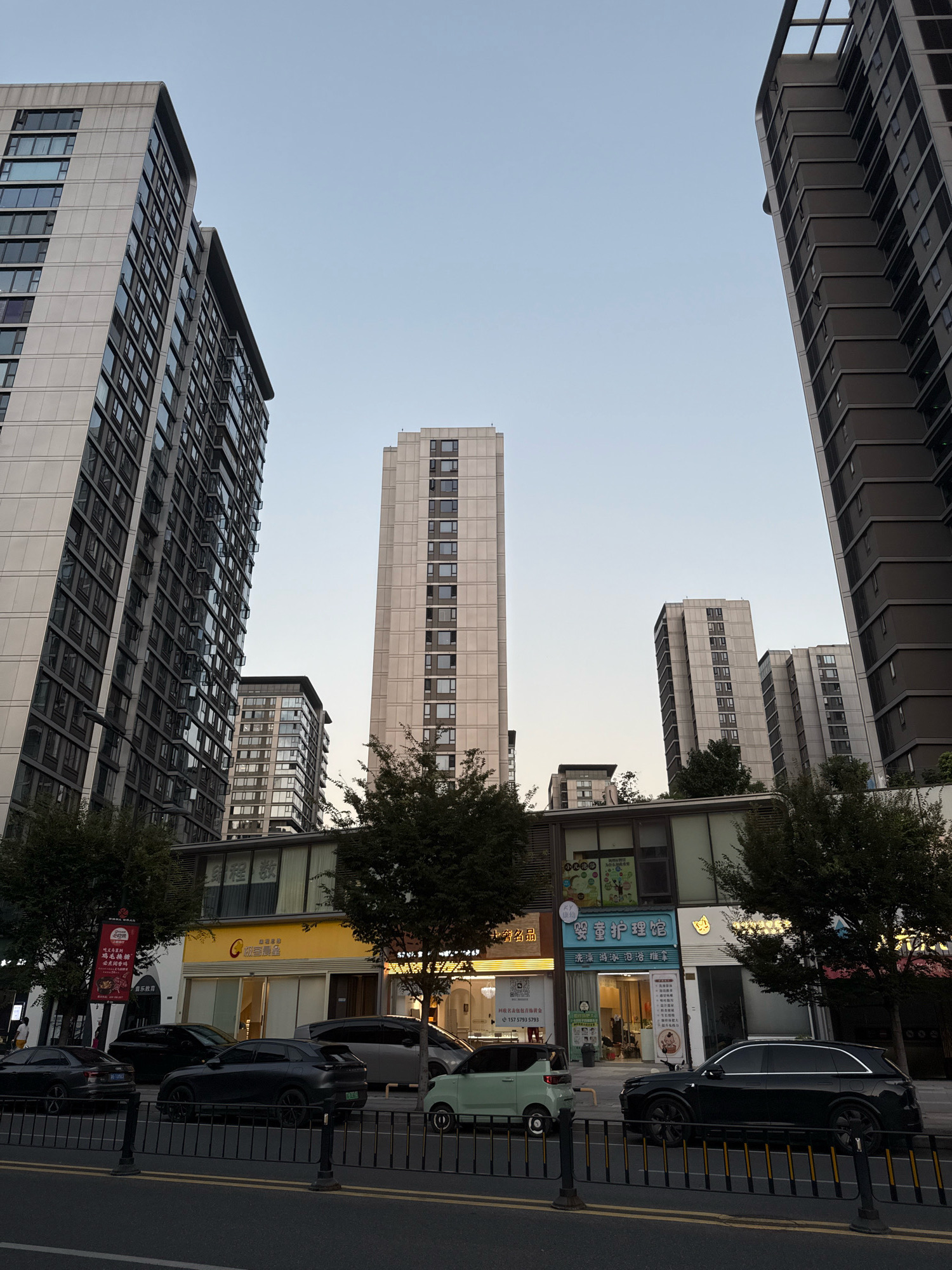 The image depicts a street scene in Yiwu, China, characterized by a clear sky and modern high-rise buildings. The foreground features a road with several dark-colored cars parked along the sidewalk, separated by a black metal fence. Behind the cars, a row of businesses with bright signage occupies the ground floor of lower buildings. These businesses include establishments with yellow, white, and blue facades, displaying various advertisements and signage, some with Chinese characters. Trees are interspersed along the street, providing some greenery. In the background, several tall residential buildings dominate the skyline, displaying a pattern of numerous windows. The overall impression is of an urban environment with a mix of residential and commercial spaces.