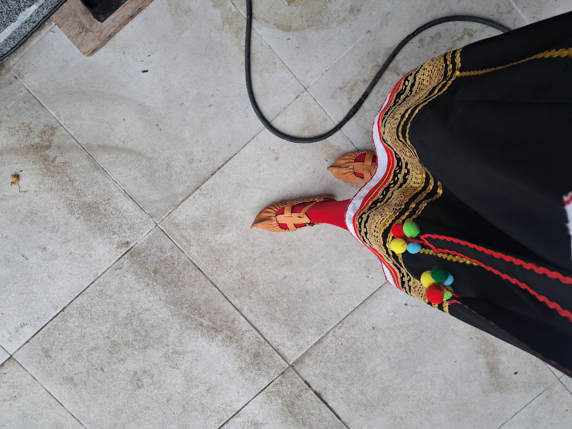 The image shows a person standing on a tiled floor in what appears to be an indoor or covered outdoor space. The tiles are light gray and show some wear. The person is wearing a traditional garment consisting of red leggings, leather sandals, and a black robe decorated with intricate gold embroidery, white and red stripes, colorful pom-poms, and red decorative stitching. A black electrical cord is visible in the background, curving across the floor near the person. A wooden pallet rests in the upper-left corner of the image. The location is Dragoman, Bulgaria.