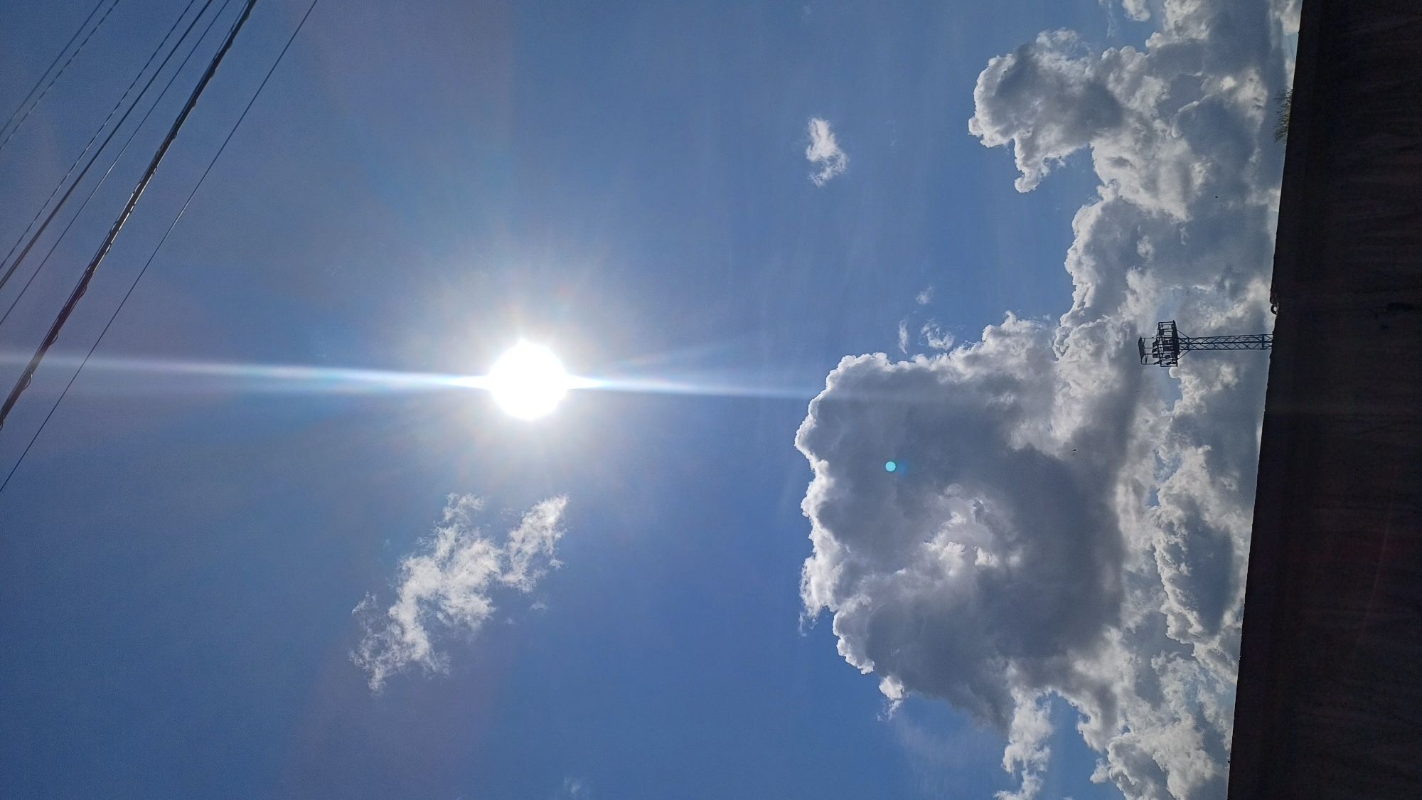 A bright sun shines in a clear blue sky, with a few scattered clouds. A tall communications tower stands silhouetted against the clouds, and a dark, low wall runs across the bottom of the frame. Overhead, power lines are visible.