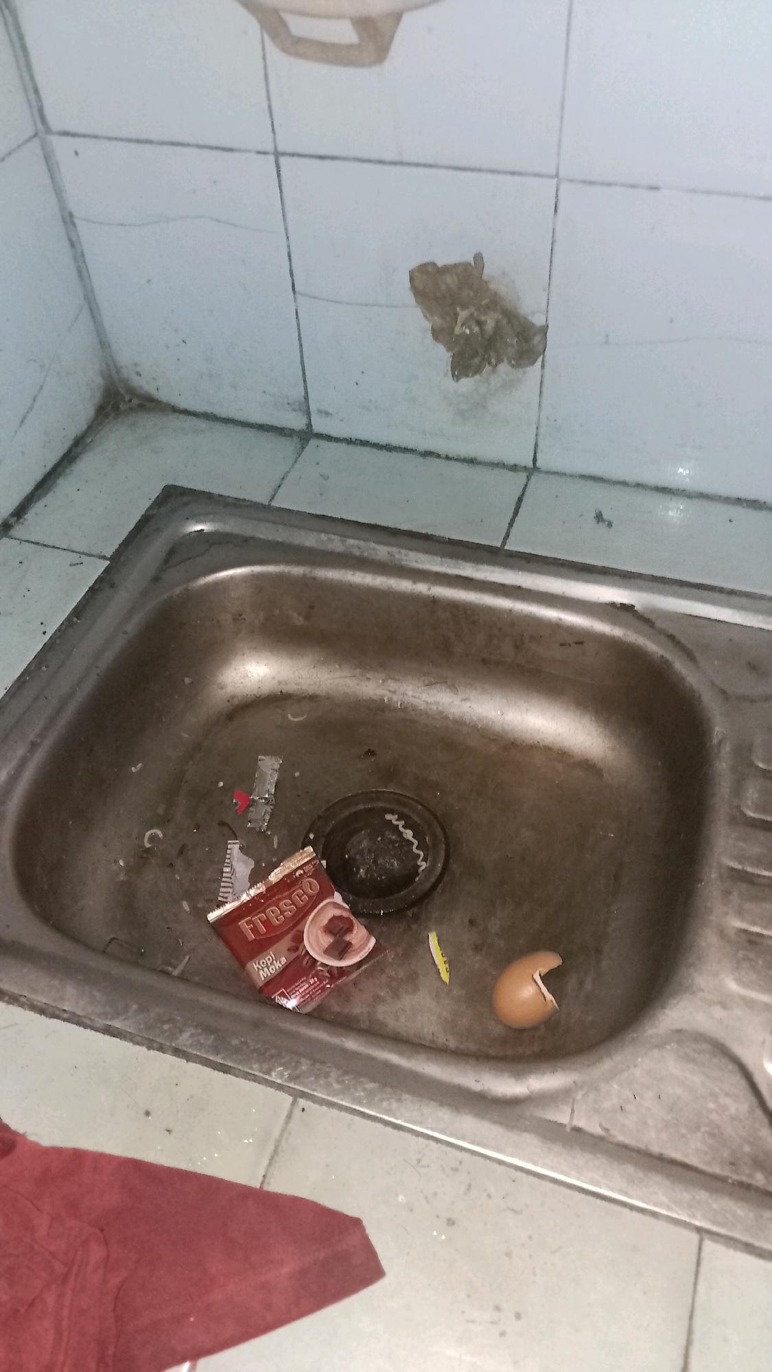The image shows a close-up, overhead view of a dirty, stainless steel sink with a tiled wall in the background. Debris is scattered throughout the sink basin, including a partially opened packet of "Fresco Kopi Moka" instant coffee, a broken eggshell, and various small pieces of trash and packaging. The tiled wall behind the sink appears grimy and has a dark stain. To the left of the sink, a portion of a red cloth or garment is visible. The overall scene suggests a neglected or recently used area, possibly a kitchen or utility sink, within a building in Mulyo Haji, Indonesia.