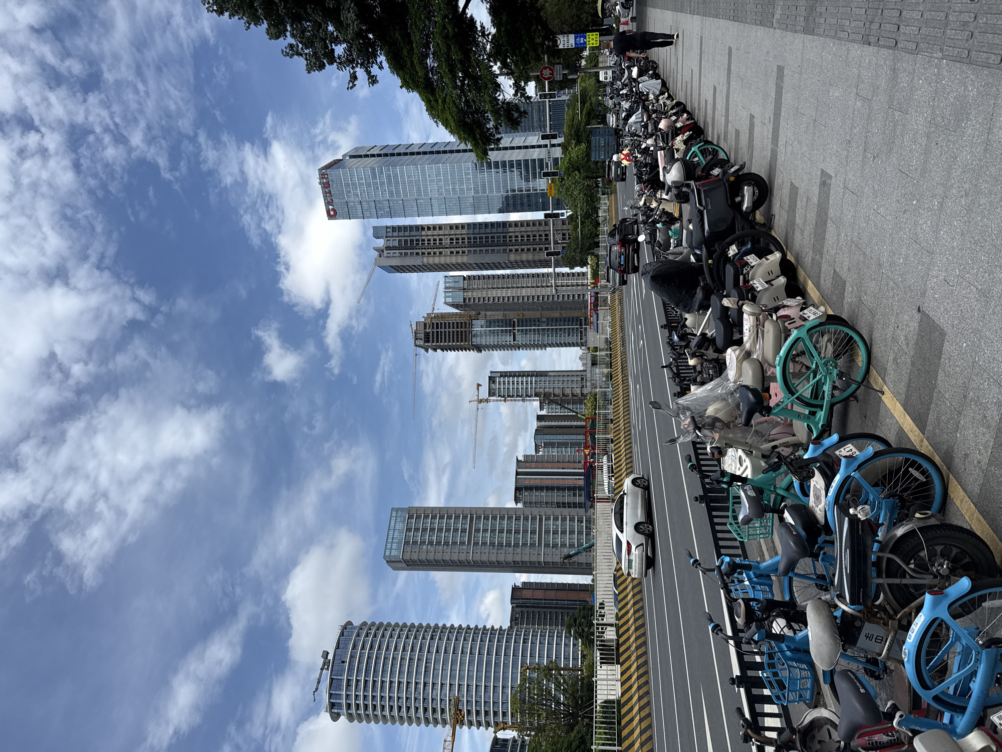 In Tianhe District, China, a street scene unfolds under a cloudy sky. A white sedan travels on an elevated highway in the background, adjacent to a sidewalk lined with numerous parked motorcycles and electric bikes. The vehicles are densely packed, with many covered by tarps. The environment features modern skyscrapers and construction cranes. A man in a black t-shirt and shorts walks on the sidewalk towards the right side of the frame. The sidewalk itself is paved with granite tiles and bordered by a textured walkway.