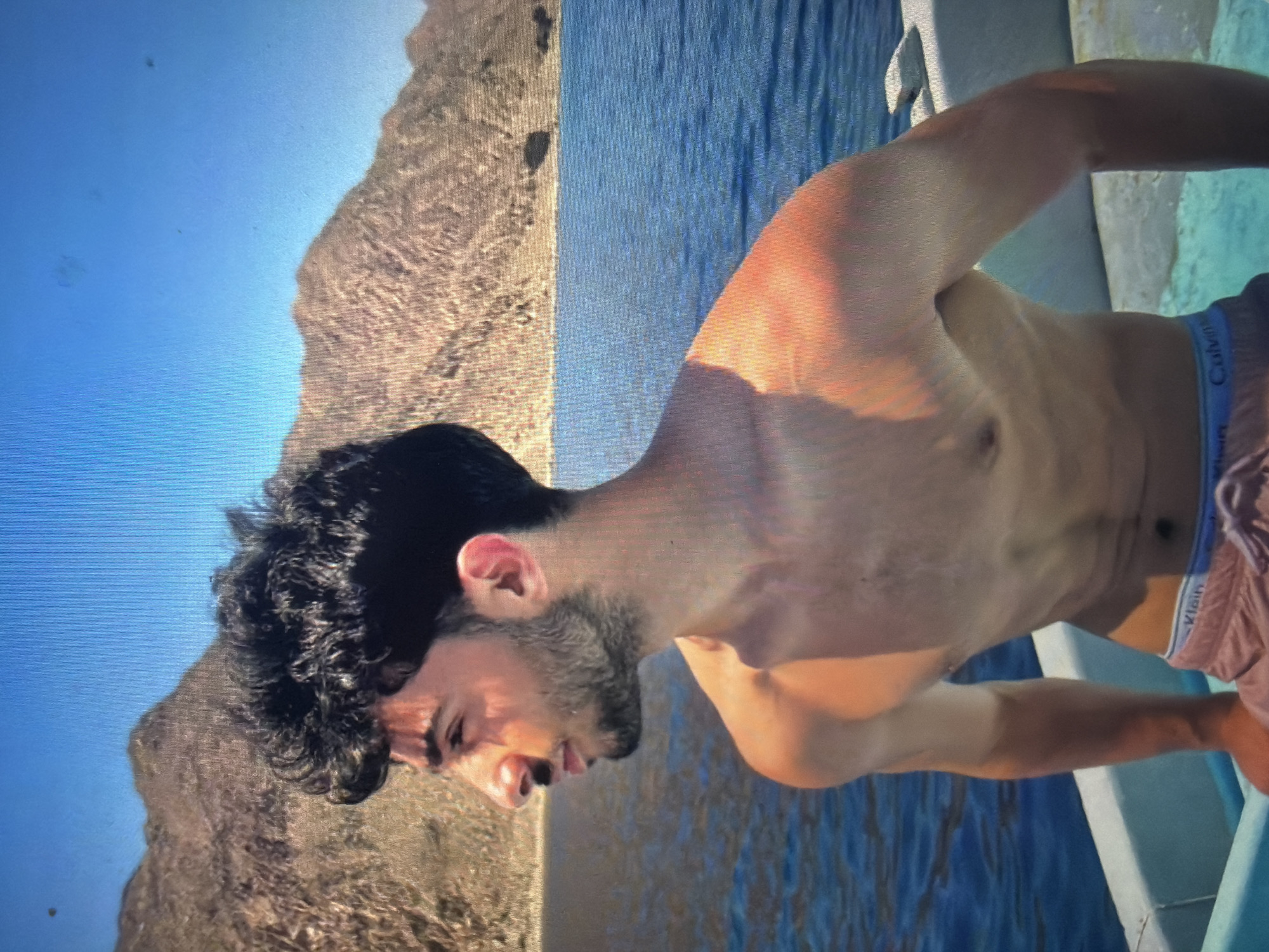 The image captures a male individual, appearing to be in his late teens or early twenties, standing or leaning within a boat. He is depicted in left profile, shirtless, revealing a tan complexion and athletic build. He has dark, curly hair, a short beard, and a mustache. He wears light-colored shorts with a visible blue waistband bearing the "Calvin Klein" brand name. The background features a large body of blue water, which extends to an opposite shore characterized by arid, rocky mountains or hills under bright sunlight. The sky above is clear and blue. Multiple small, dark, irregular marks, consistent with dust or sensor spots, are visible against the bright blue sky. This scene is located in the city of Shumen, Bulgaria.