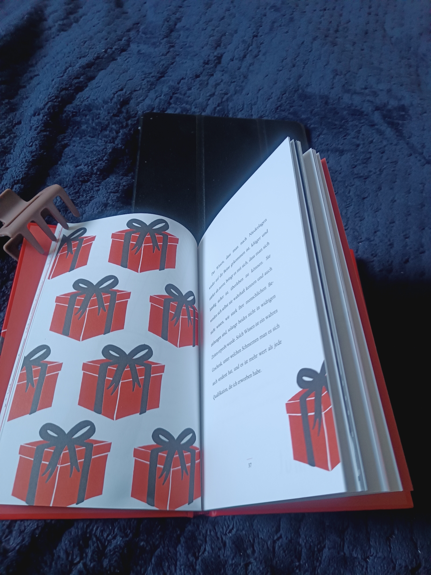 The image shows an open book with red gift box illustrations on the left page and text on the right page. The book is lying on a dark blue, textured surface, possibly a blanket or carpet. A dark-colored tablet device is visible behind the book. A brown hair clip is positioned on the left side, holding open the left page of the book. The text on the right page appears to be in German and includes the number "37" at the bottom left. There is no indication of the city of Weilburg, Germany, in the image or its context.