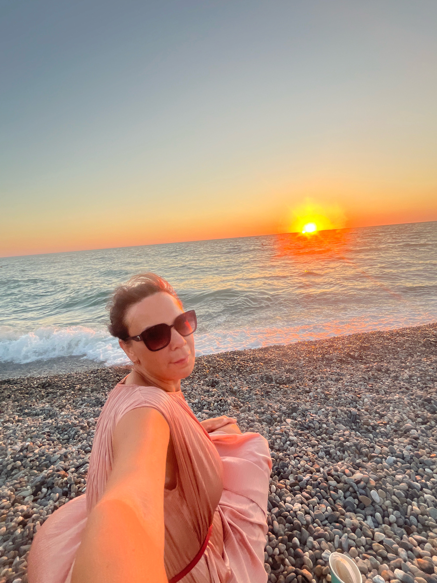 A woman is taking a selfie on a pebble beach in Sochi, Russia. She is wearing sunglasses and a pink dress, and is sitting on the beach. The sun is setting over the sea, casting a warm glow on the scene. The waves are gently rolling onto the shore. There is a disposable cup in the lower right corner of the image.