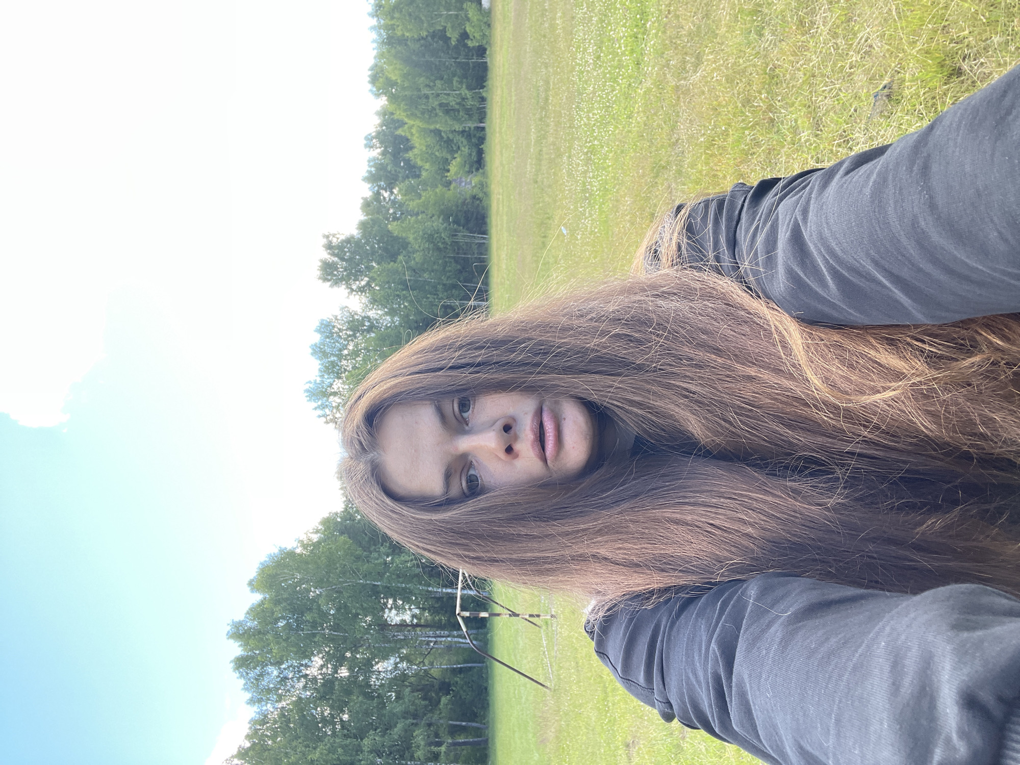 This media file is a portrait-style photograph of a young person outdoors. The subject, with long, light brown hair and fair skin, is positioned in the foreground, with their face looking directly at the camera. They are wearing a dark long-sleeved shirt and dark pants. The subject's hair is a prominent feature, covering a large portion of the frame and framing their face.
The background suggests a natural, outdoor setting. A grassy field with scattered white flowers extends into the distance, bordered by a line of trees. In the middle ground, a partially visible soccer goal can be discerned, implying the location might be a sports field or park. The sky is bright and appears to be daytime, with a few wispy clouds visible, suggesting fair weather. The orientation of the photograph is such that the subject appears to be lying down or the camera is held at an angle.
The overall mood is casual and relaxed, with the subject making direct eye contact with the viewer. There is no visible text in the image. The location context provided is Заречный, Russia.