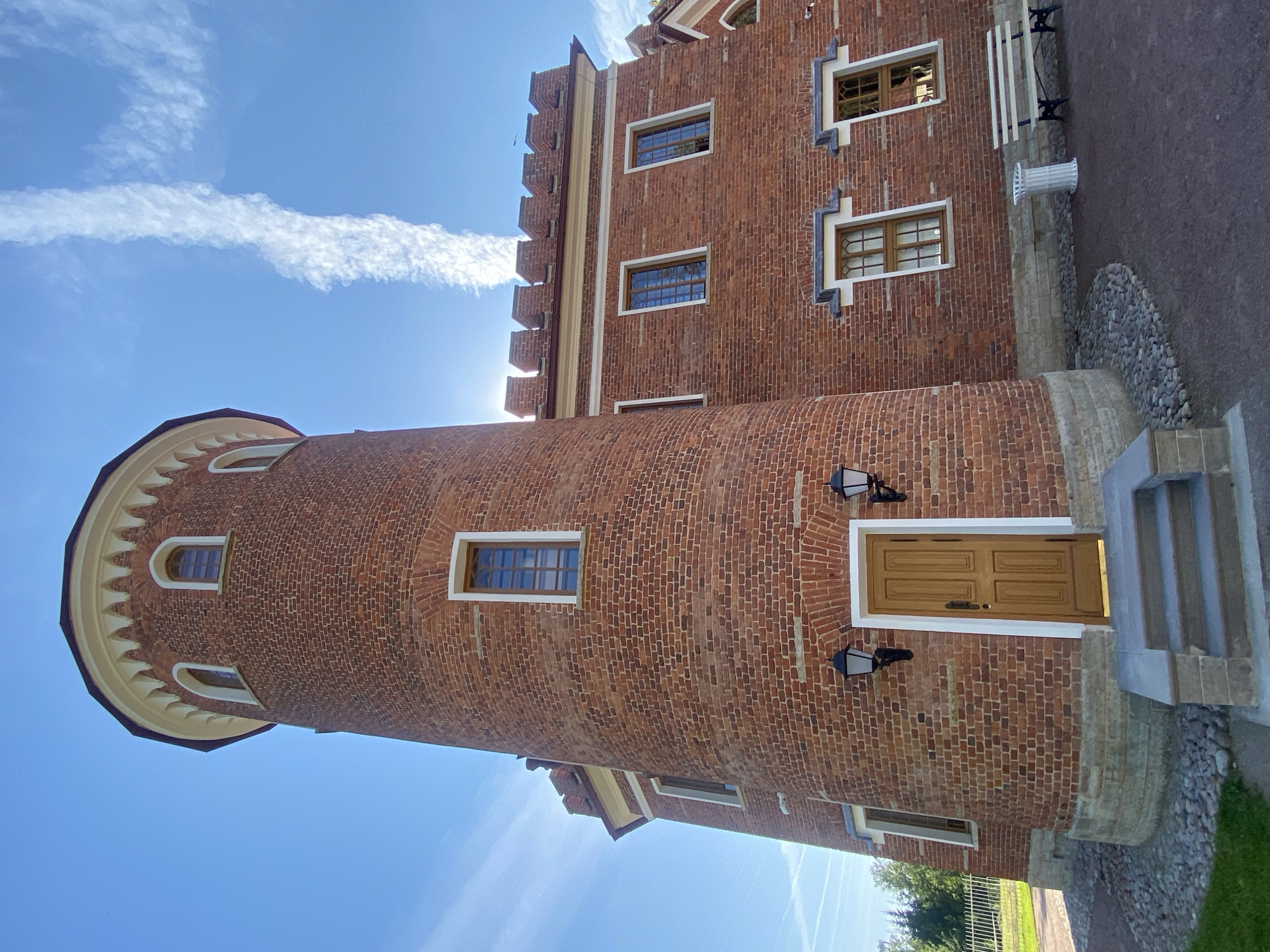 The image shows a red brick tower and adjacent building with a crenellated roofline under a bright blue sky with scattered white clouds. The architecture suggests a historical or whimsical design, reminiscent of a castle or folly. The tower has arched windows and a rounded base. A wooden door with a dark handle is set into the tower, approached by a set of stone steps. Two black lanterns hang on either side of the door. The building to the right has rectangular windows with intricate wooden frames. A white bench and a white cylindrical trash receptacle are placed on the ground in front of this building. The surrounding area is paved with dark gravel and cobblestones. A white metal fence is visible in the background, with trees beyond it. The location is identified as the "Alexandrovsky Park," within the city of Pushkin, Russia. There are no people visible in the image.