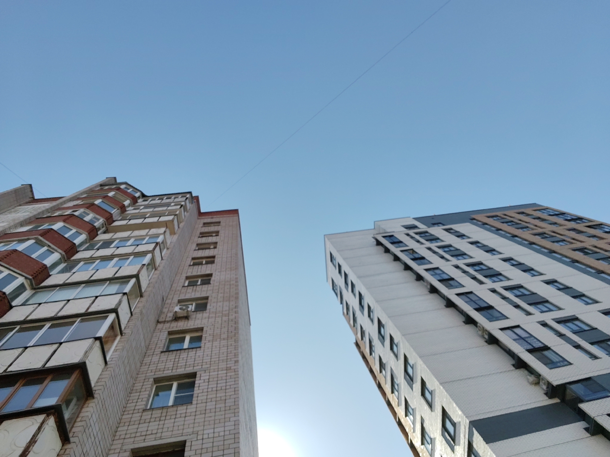 The image depicts two tall buildings against a clear blue sky. The building on the left is constructed of light beige brick with protruding balconies and red brick accents near the top. The building on the right has a white facade with grey and tan accents, and many rectangular windows. There is a thin wire stretching across the sky between the two buildings. The perspective is looking upwards at the buildings, suggesting the photo was taken from street level. While I am unable to confirm the location is in Izhevsk, Russia, the architectural style is consistent with buildings found in many post-Soviet cities.