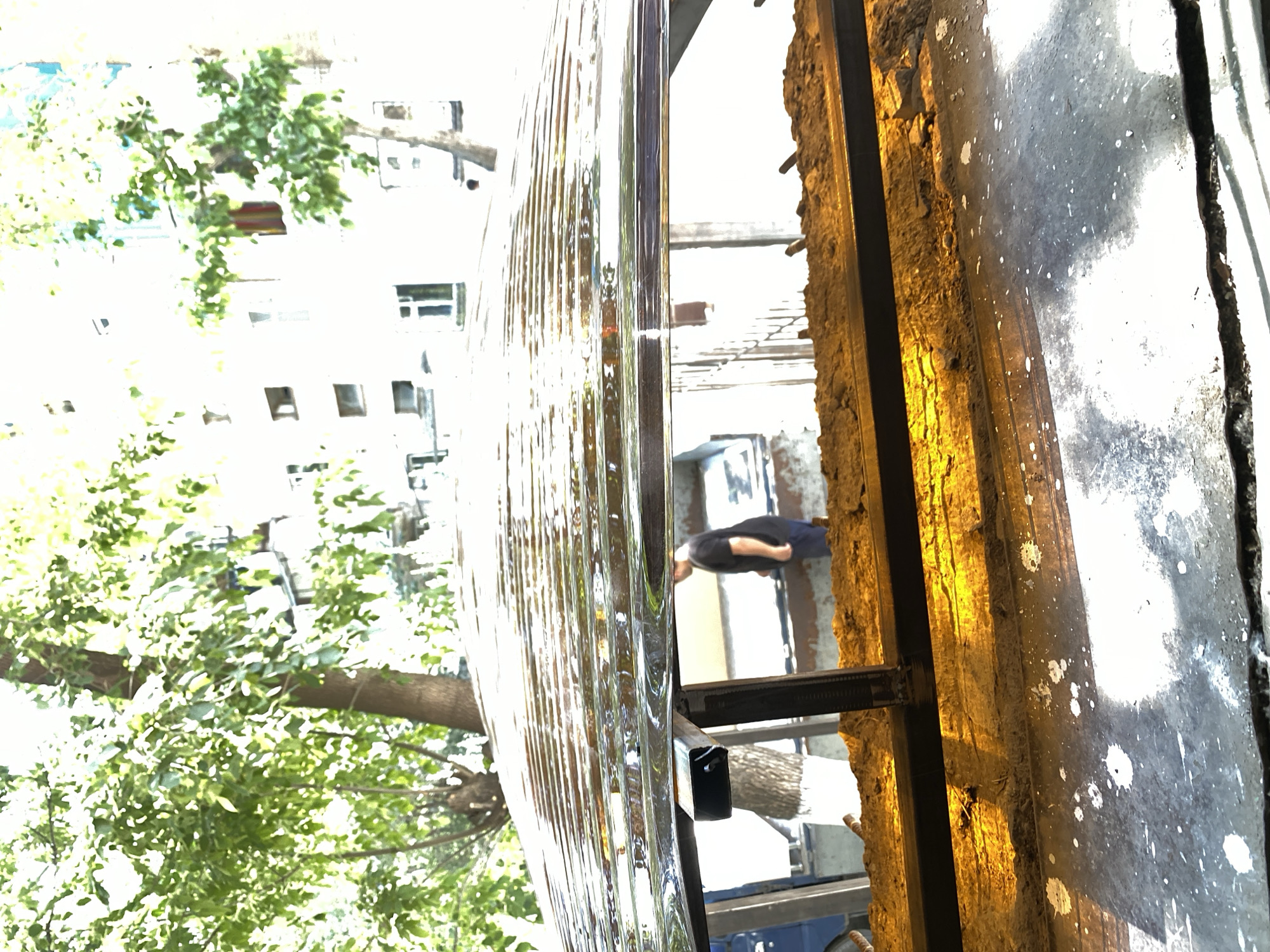 The image shows a view from under a corrugated metal roof, looking out towards a street scene. The roof has water droplets on it, creating a distorted reflection of the environment. In the background, a person wearing a dark t-shirt and pants is standing on a sidewalk next to a building. Trees with green foliage are visible in the foreground and background, suggesting an outdoor setting in a city. The building appears to be multi-story with windows. The overall scene suggests an urban environment, possibly during or after rain. The location is identified as Manas, Kyrgyzstan.