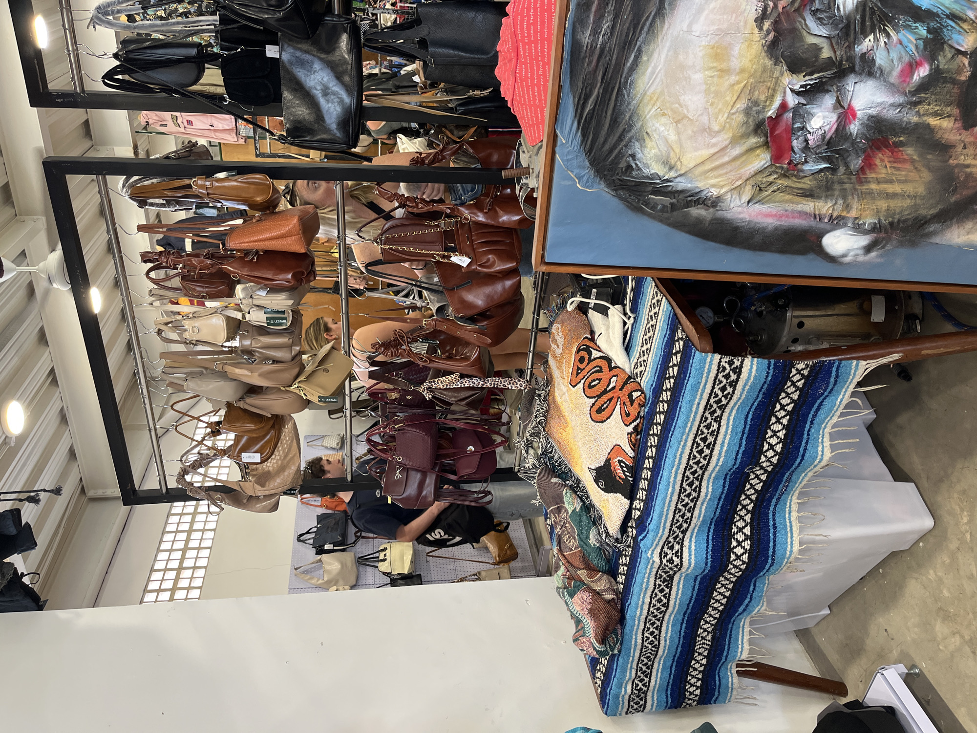 An indoor market stall in North Kuta, Indonesia displays an assortment of leather handbags hanging from racks. A table in the foreground is draped with a blue and white striped blanket, on which several woven textiles and pillows are arranged. To the right, a portion of a painting depicting a person's face is visible, with bold, abstract brushstrokes. In the background, a man wearing a dark t-shirt and jeans stands near a display of bags, looking at the merchandise. The market has a bright, overhead lighting system and exposed ceiling beams.