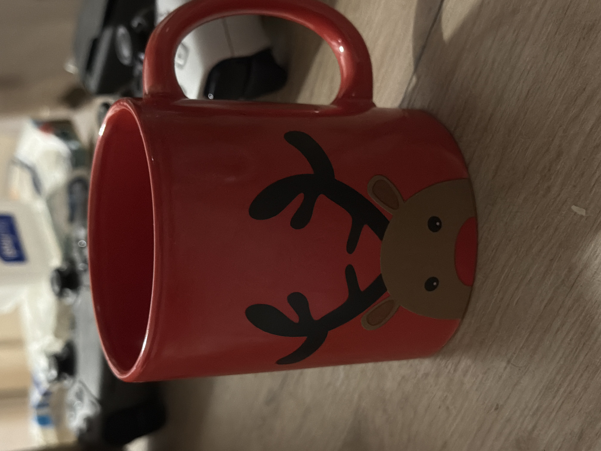 The image shows a red ceramic mug with a reindeer design, featuring black antlers, brown face, and a red nose, resting on a light brown surface, possibly a wooden table. In the background, partially visible, are blurred objects that appear to be kitchen appliances and packaged goods. The location in Podgorica, Montenegro, is not discernible from the given visual information.
