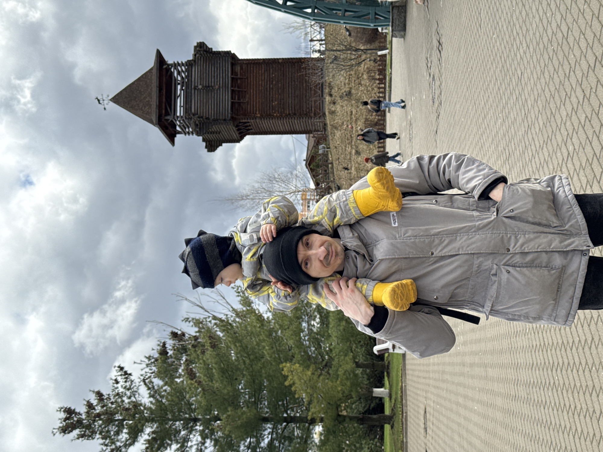 A father carries his son on his shoulders in a park. The father, wearing a black beanie and a light grey jacket, is holding the son's legs, which are clad in bright yellow boots. The son, wearing a striped dinosaur-themed beanie and a grey jacket, appears to be looking at something to the left. In the background, there's a wooden tower structure against a cloudy sky. Other individuals are walking in the background on a brick-paved area. The location is in Izhevsk, Russia.