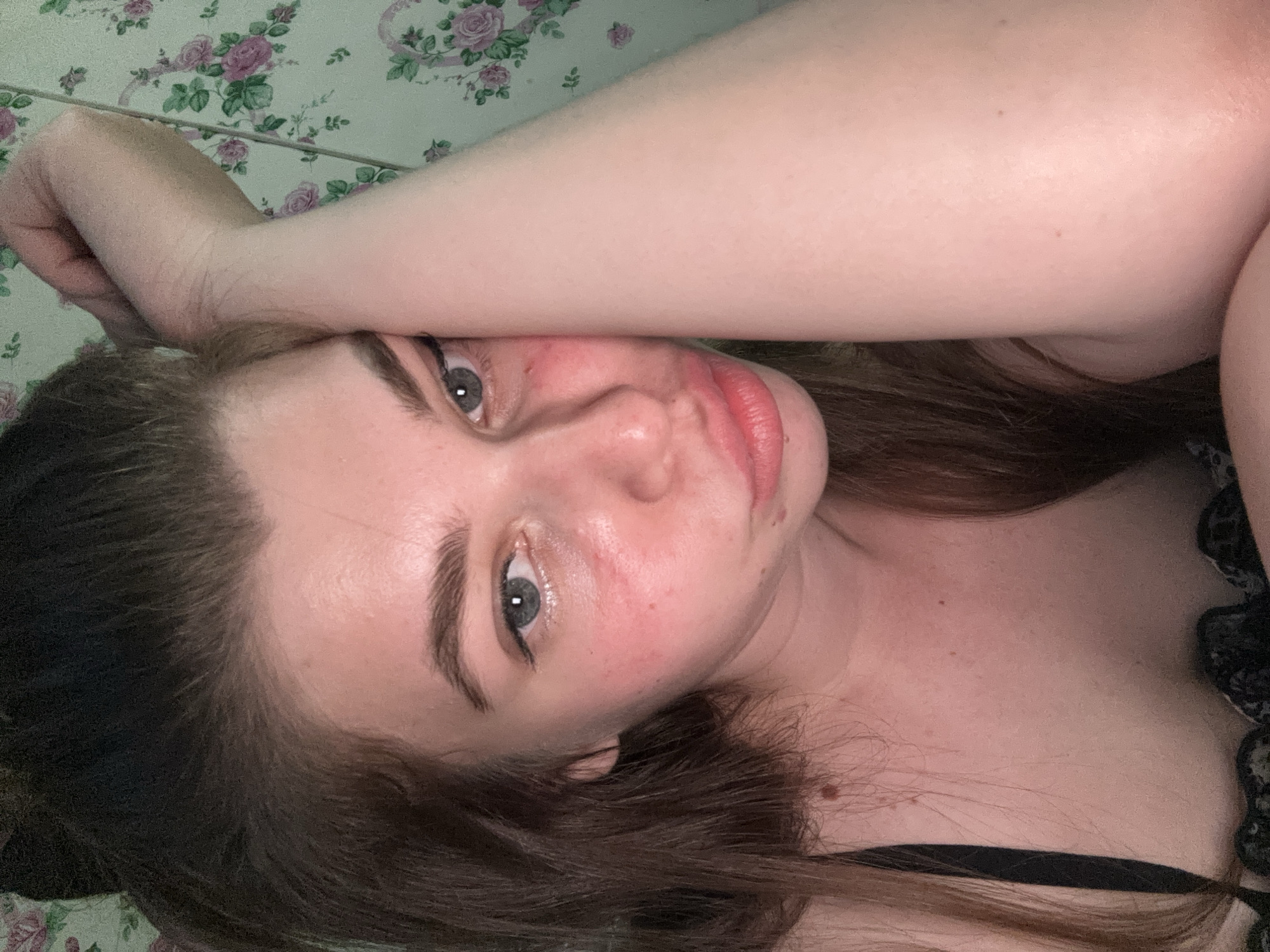 A young woman lies on a floral patterned surface, likely a bed or blanket. Her left arm is raised and bent, resting near her forehead. She has fair skin, brown hair, and light-colored eyes with dark eyeliner. She is wearing a black top. The scene appears to be indoors. There is no information about the location of the city of Мещёры, Russia.