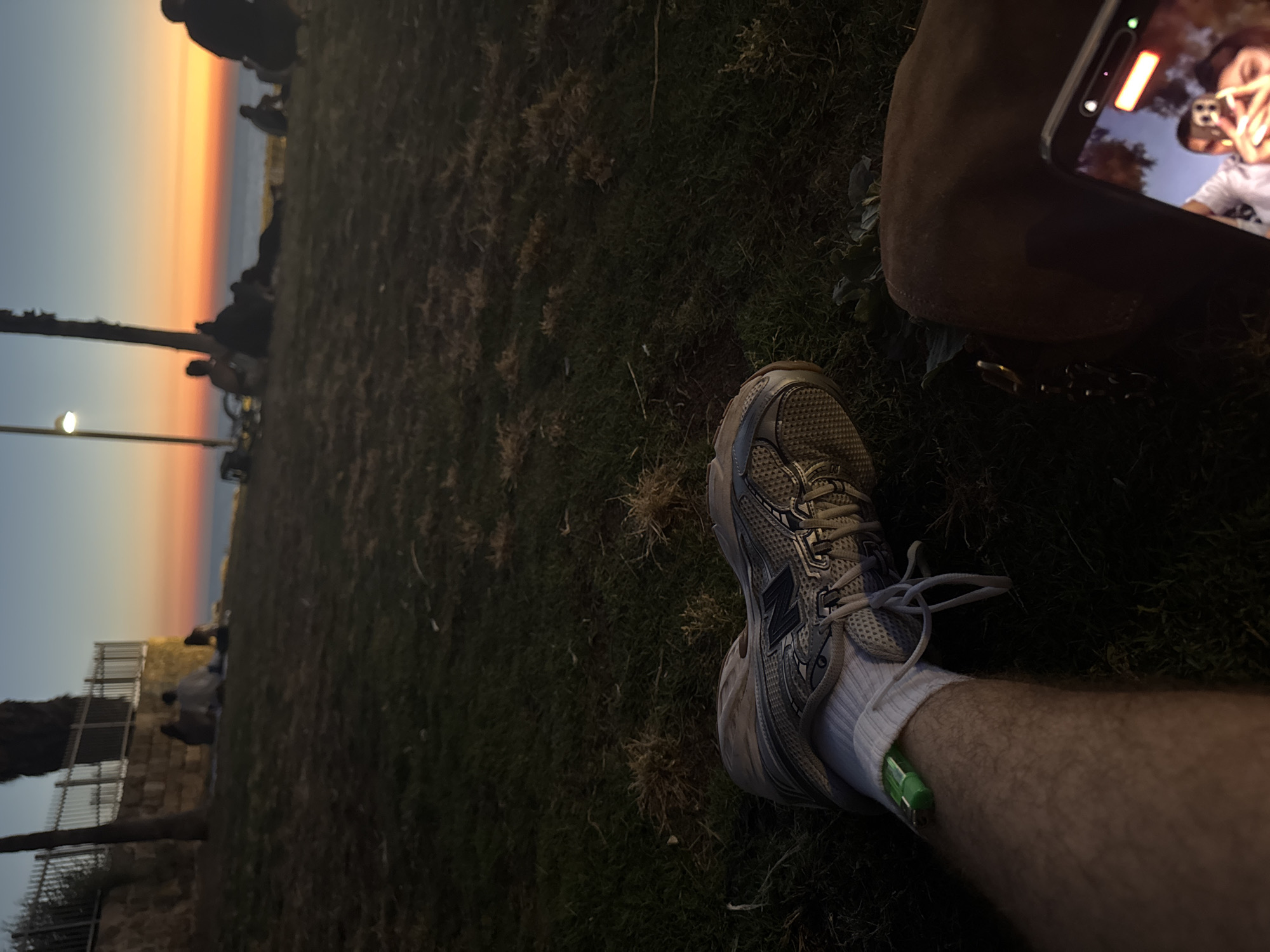 The image captures a low-angle view of a person sitting on a grassy slope, looking out towards a sunset over the Mediterranean Sea. The sky displays a gradient of orange, yellow, and blue, indicative of dusk. In the foreground, the person's leg is visible, wearing a white sock and a grey athletic shoe. A green lighter is tucked into the sock. A dark brown fabric, likely pants or a blanket, is also in the frame. In the background, silhouetted figures are scattered across the grassy area and along the coastline. Some are sitting, while others appear to be walking or standing. A metal fence and stone wall are situated further up the slope. The overall scene suggests a relaxed evening gathering at a public park in Tel-Aviv, Israel, possibly near the coastline, enjoying the sunset.