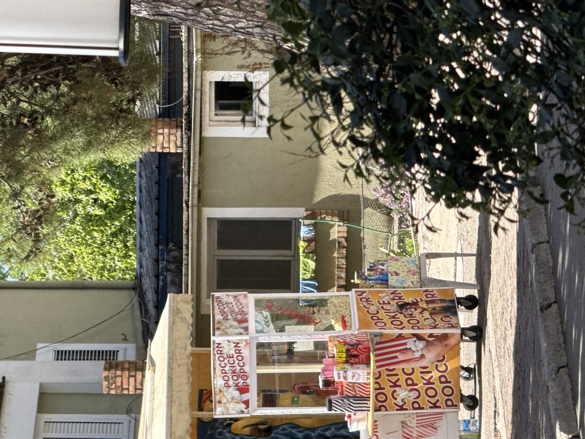 Here is a description of the image:
The image shows a popcorn kiosk located on a street, likely in Budva, Montenegro. The kiosk is white and features transparent display cases, displaying a variety of popcorn and other snacks. The kiosk advertises "POPCORN" and "KOKICE" in English and presumably Montenegrin, respectively. The background reveals a street with buildings, including one with windows and clothing hanging to dry. Vegetation is present with trees and plants in the foreground and background.