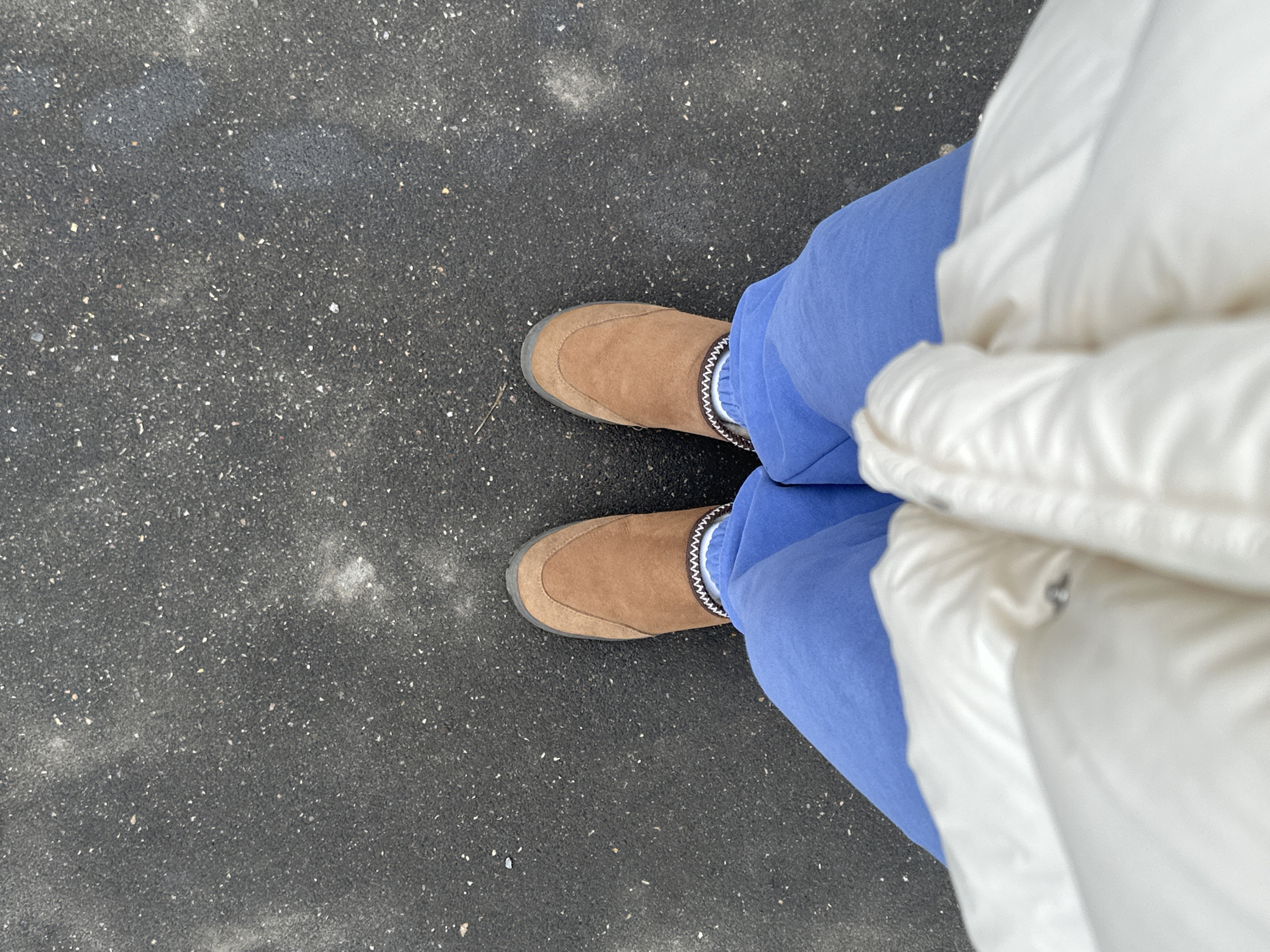 A first-person perspective photograph shows a person standing on a gray asphalt surface. The person is wearing brown slip-on shoes with a decorative trim, light blue sweatpants, and a white puffer jacket. The location is not identifiable as Krasnogorsk, Russia, based on the visual information provided.