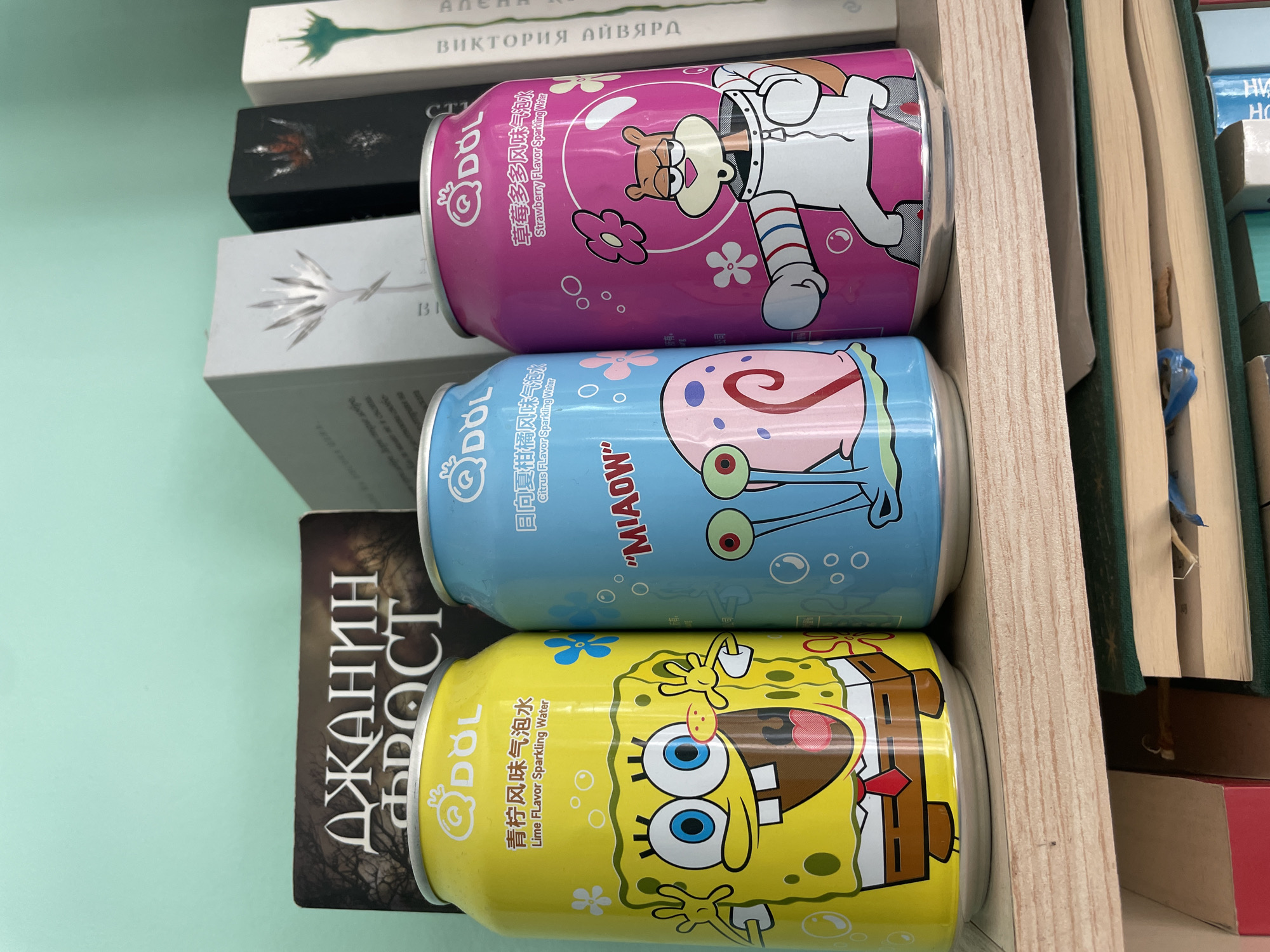 An eye-level shot captures three brightly colored beverage cans featuring Spongebob Squarepants characters, arranged on a light-colored wooden shelf. The cans are positioned against a light teal-green wall, with several books partially visible behind and beneath them.
From left to right:
1. A vibrant yellow can features a smiling Spongebob Squarepants. The text "Q DOL" is prominently displayed at the top, followed by "青柠风味气泡水" (Lime Flavor Sparkling Water) in both Chinese and English.
2. A medium-blue can showcases Gary the Snail, with the word "MIAOW" written in red capital letters. Above Gary, "Q DOL" is visible, along with "日向夏橘风味气泡水" (Citrus Flavor Sparkling Water) in Chinese and English.
3. A bright pink can depicts Sandy Cheeks in her space suit. "Q DOL" is at the top, followed by "草莓多风味气泡水" (Strawberry Flavor Sparkling Water) in Chinese and English.
Behind the cans, several books stand upright. The leftmost visible book, behind the yellow can, has a dark cover featuring bare trees and Cyrillic text reading "ДЖАНИН ФРОСТ" (Janine Frost). To the right, other books have more minimalist designs, including white spines with stylized silver, black spines with red, and white spines with green motifs. Some Cyrillic text, such as "ВИКТОРИЯ ЛЮБЭРА" (Victoria Lyubera), is partially visible on the spine of one of these books.
The wooden shelf beneath the cans exhibits a distinct grain pattern. Below this shelf, additional books with green and blue spines are stacked, partially obscured. The overall lighting is even, suggesting an indoor setting in Unknown, Unknown.