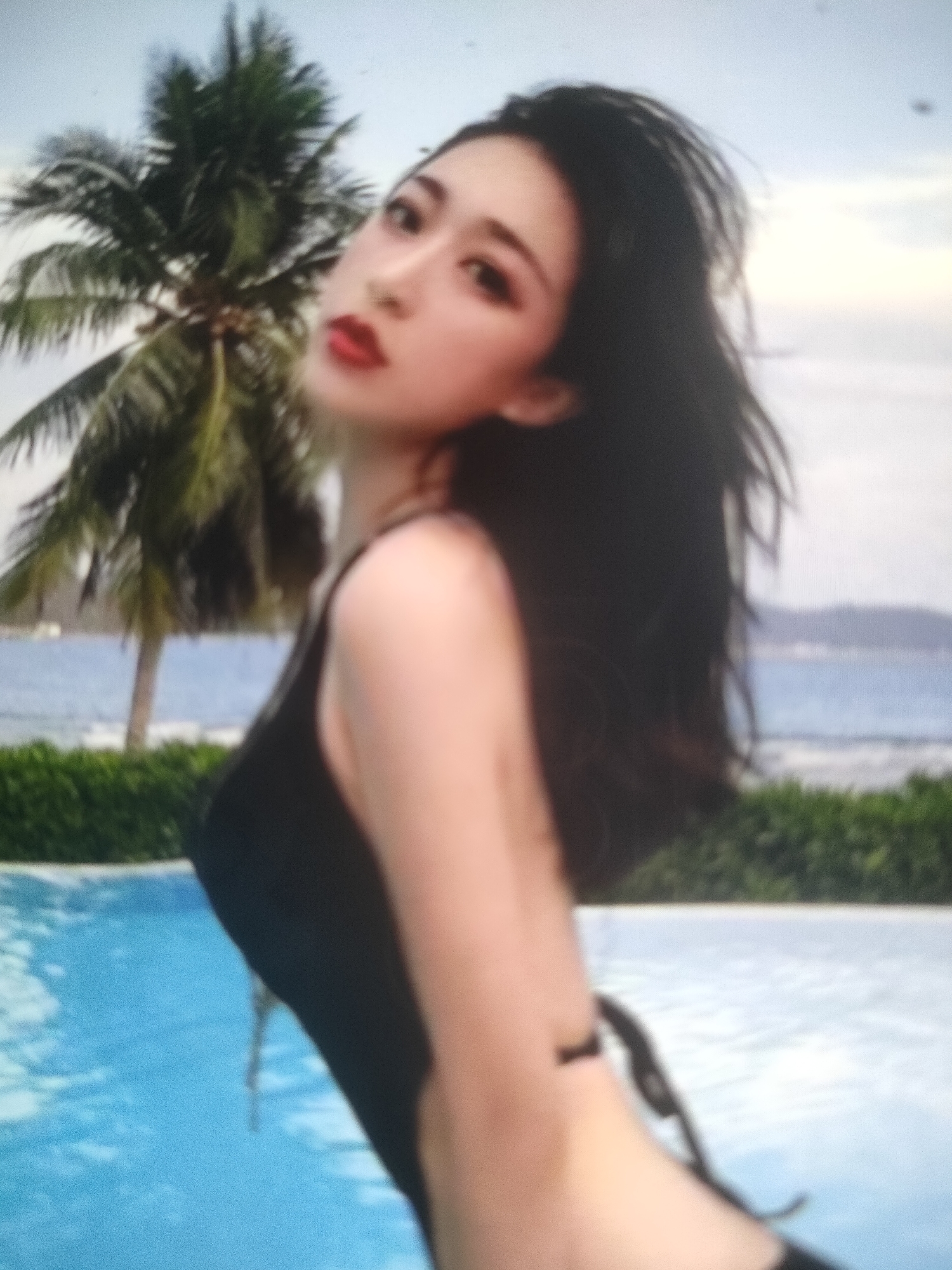 The image shows a blurry, close-up view of a woman in a black swimsuit. She is positioned to the right side of the frame, with her head turned slightly away from the camera and her gaze directed upwards. Her long, dark hair flows over her shoulder. The background is out of focus but appears to be a tropical setting with a palm tree, the ocean, and lush green foliage. The water in the foreground is a bright blue, suggesting a swimming pool. The overall scene evokes a relaxed, vacation-like atmosphere, possibly at a beach resort or a private villa. The image quality is low, with a grainy texture and soft focus, which obscures finer details of the woman and her surroundings. There is no indication of the specific location within Padang Dalom, Indonesia.