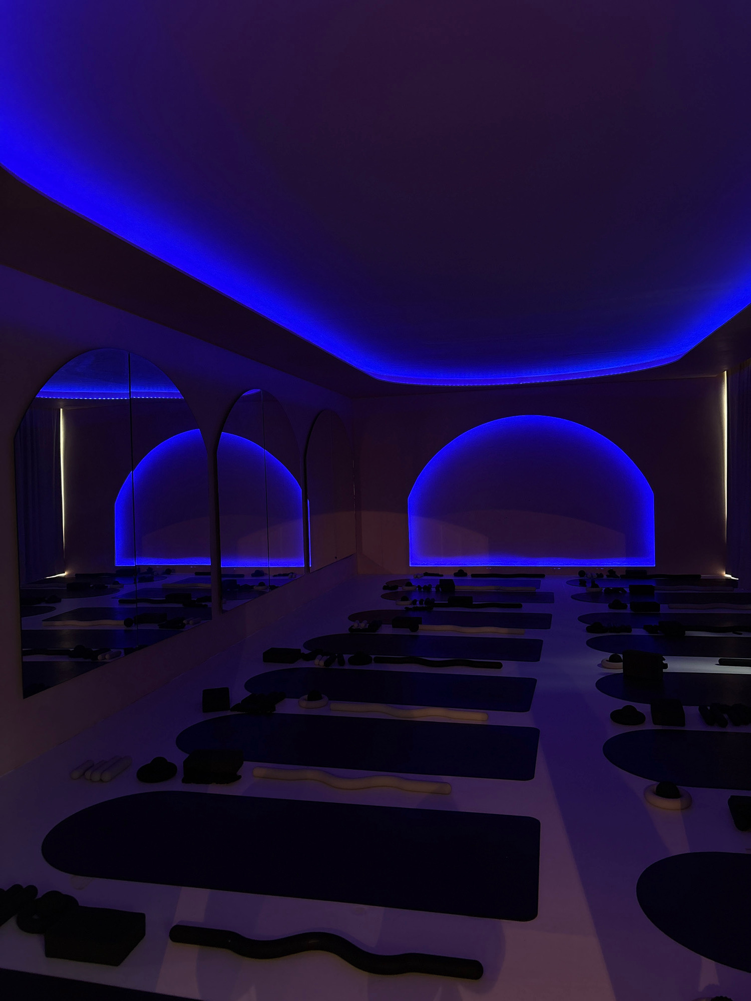 This image depicts an empty, dimly lit yoga studio with a modern, minimalist aesthetic. The primary illumination comes from vibrant blue LED lights embedded along the ceiling's curved edges and a large, arch-shaped light feature on the back wall, creating a moody and atmospheric ambiance. The floor is a white or light-colored surface, overlaid with dark yoga mats arranged in neat rows, suggesting preparation for a class. Various yoga props, such as foam rollers and blocks, are scattered on the floor in front of the mats. The studio features large arched mirrors along one wall, reflecting the space and the blue lighting. The overall impression is one of a serene and technologically advanced wellness environment. There are no people present in the image. The location is confirmed to be in Mexico City, Mexico.