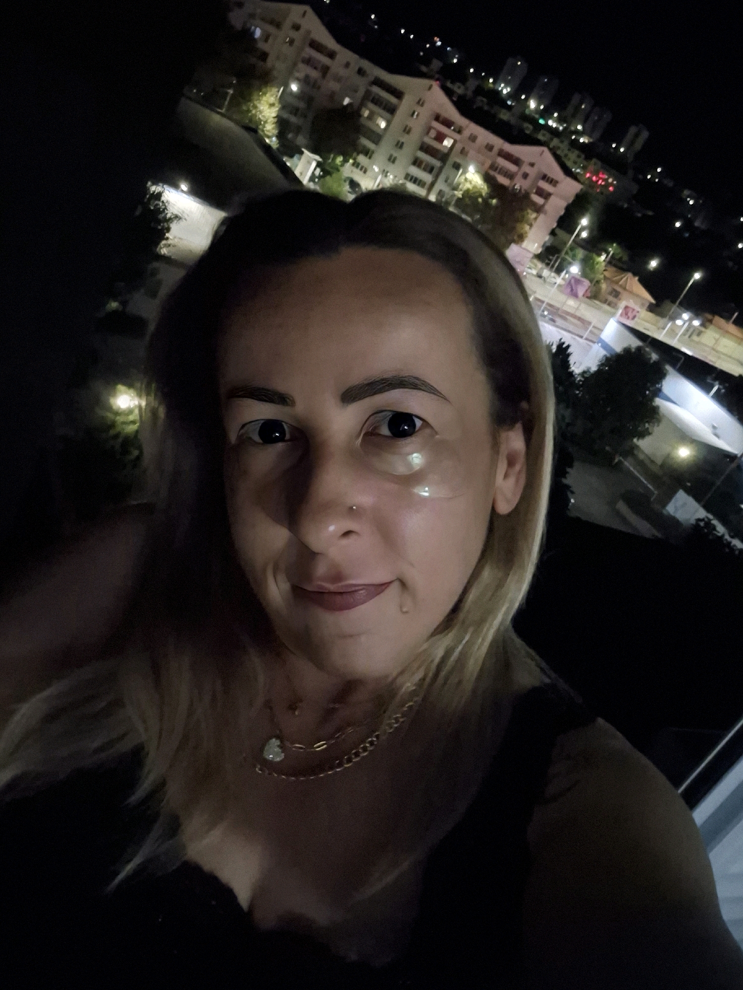A woman takes a selfie at night with an urban landscape in the background. She has blonde hair, dark eyebrows, and is wearing a black top and two necklaces. Her eyes are dark and she has clear patches under her eyes. The background consists of buildings with lights on, streetlights, and trees. The overall lighting suggests it is nighttime.