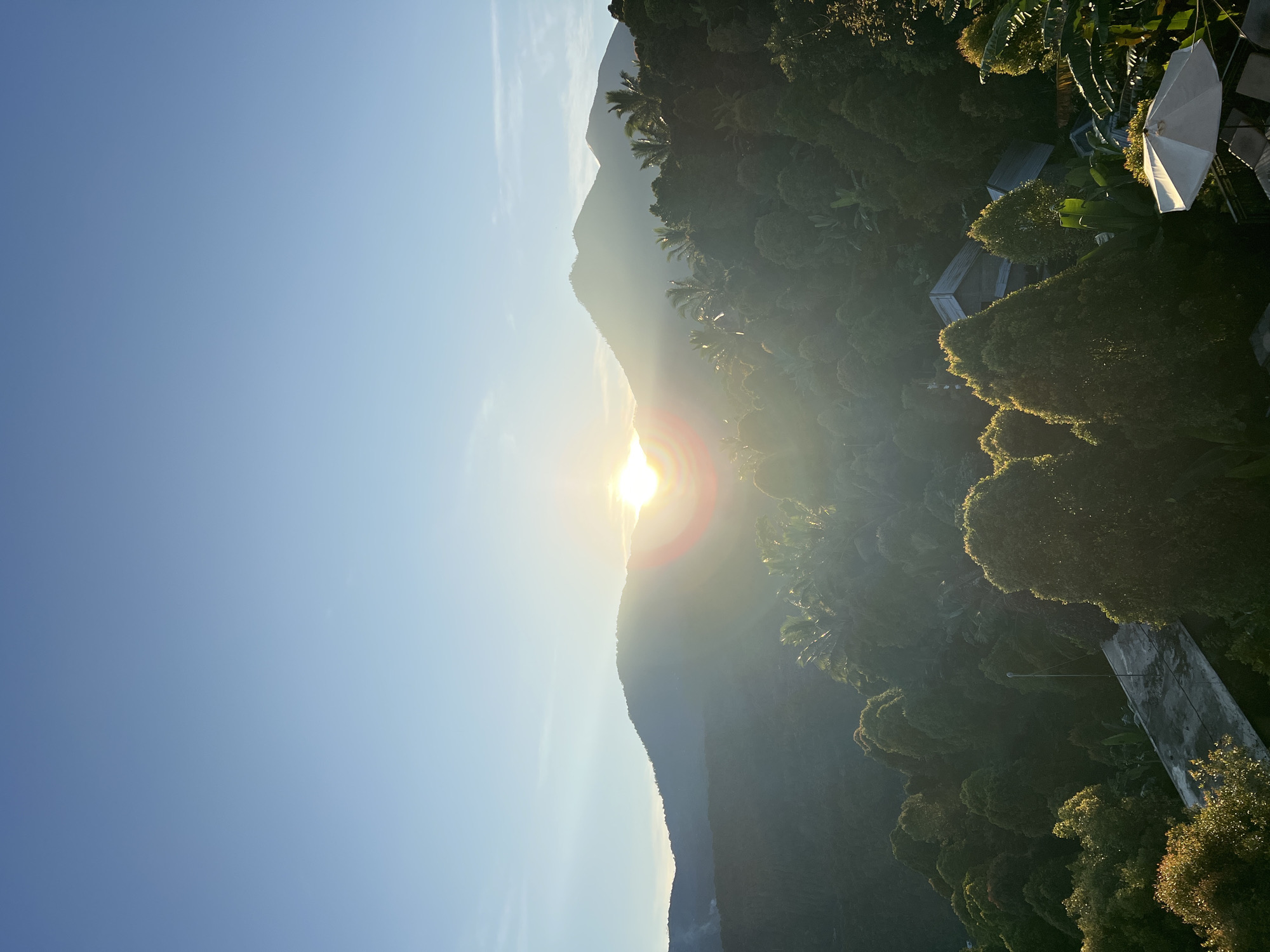 The image captures a landscape under bright sunlight. The scene is oriented vertically, showing a view of hills or mountains, partially obscuring the sun, with the sun's rays appearing to burst through the peaks. Lush, green vegetation covers the slope of the hills. A clear blue sky fills the upper portion of the image. In the right side of the image a patio umbrella, and a building with a grey roof are visible. The city of location in this image is unknown, in Indonesia.