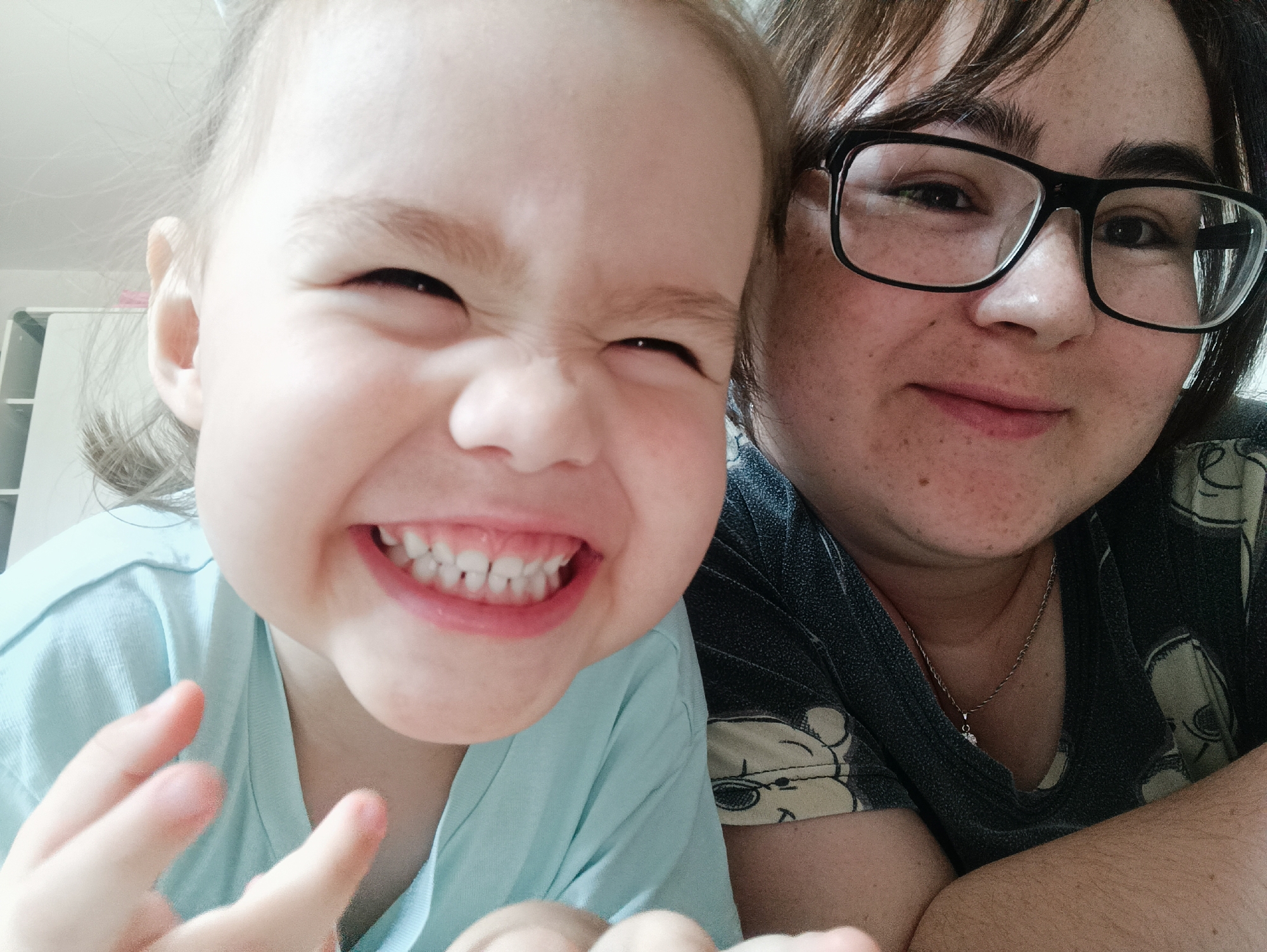 This is a close-up, indoor selfie featuring a young child and an adult. The child, positioned on the left, is looking directly at the camera with a wide, toothy grin, eyes squinted, and a playful expression. The child is wearing a light blue t-shirt and their fingers are visible in the foreground, appearing blurred from movement. To the right of the child is an adult wearing black-rimmed glasses and a dark t-shirt with a pattern of cartoon characters. The adult's expression is more subdued, with a slight smile. The background is out of focus, suggesting an indoor setting, possibly a home. There are no specific landmarks visible that would indicate the location in the city of Noviy, Russia. The lighting appears to be natural, coming from an unseen source.