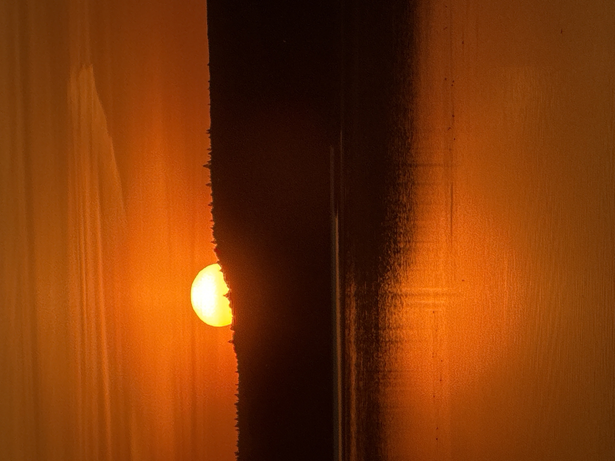 The image shows a sunset in Votkinsk, Russia. A bright, partially obscured sun is visible on the left, with a dark, vertical object partially blocking it. The surrounding environment is predominantly orange-toned, suggesting the warm light of the setting sun. The texture of the background and the dark object appears to have a grainy quality.