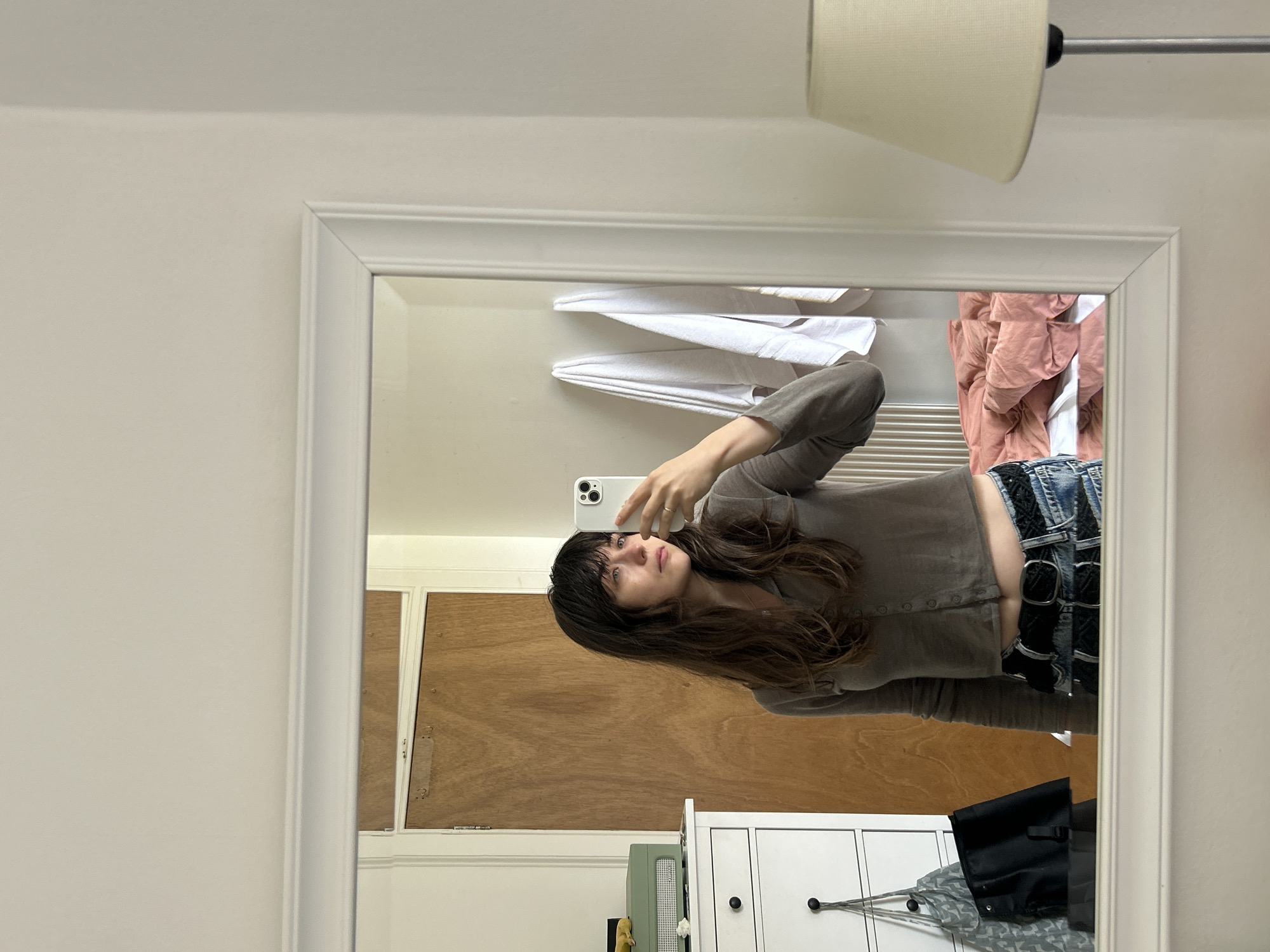 A young woman with dark, wavy hair and bangs is taking a mirror selfie in what appears to be a bedroom. She is wearing a grey, button-up, cropped long-sleeved shirt and denim shorts with a black belt. She is holding a white smartphone in her right hand, angled to capture her reflection. In the mirror, behind her, a wooden door is visible. To the left of the door, a white dresser with a green stereo system on top is present. Hanging on the dresser are two bags, one patterned and one black. To the right of the woman in the reflection are two white towels hanging on what looks like a rack. Further to the right, a corner of a bed with pink and white bedding is visible. Outside the mirror's reflection, to the right of the frame, a cream-colored lampshade of a floor lamp is visible. The overall environment suggests a casual, lived-in space. The specific location within the City of Edinburgh, United Kingdom, cannot be determined from the visual information alone.
