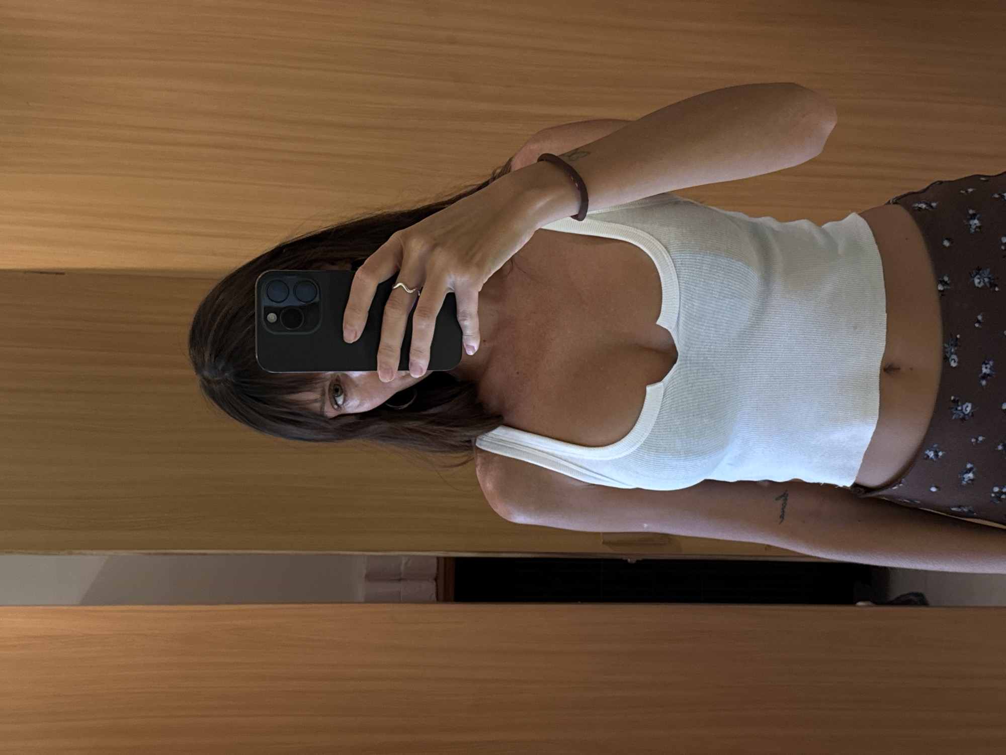A woman is taking a selfie in a room with wood-paneled walls. She is wearing a white tank top and a floral skirt. She is holding a black smartphone in her left hand. She has dark hair and light eyes. She has a ring on her left ring finger, and a bracelet on her right wrist. A tattoo is visible on her right arm. The location is likely Mengwi, Indonesia, based on the assumption that the request to include the location is related to the image.
