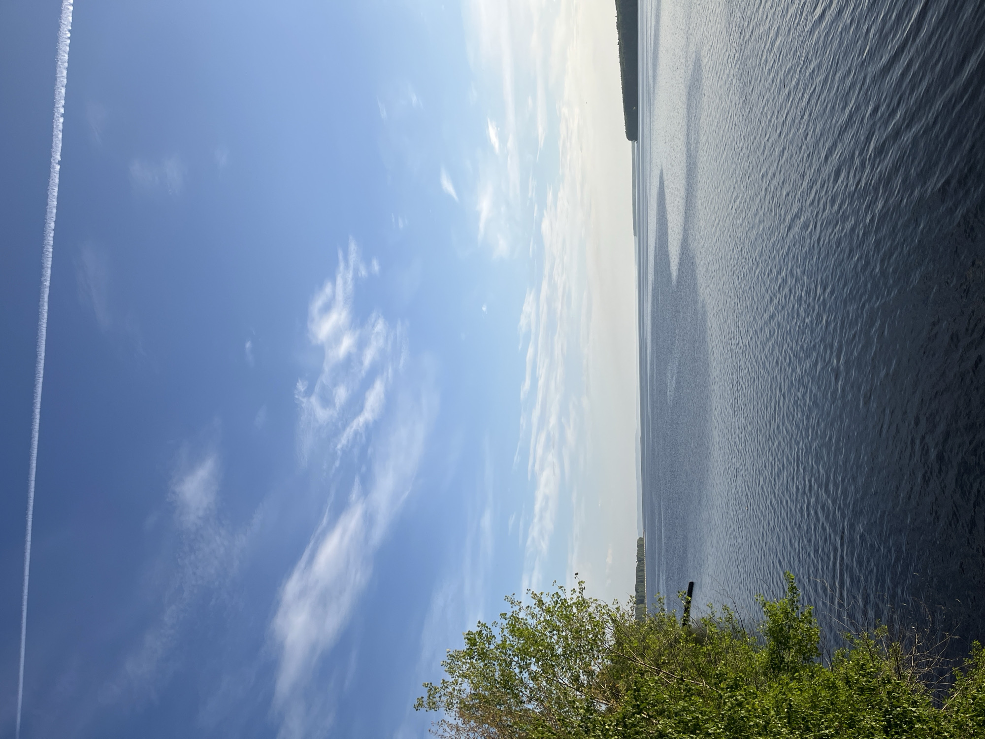 The image shows a body of water, likely the Kama River, near Chaikovsky, Russia. The sky is predominantly blue with scattered clouds and a prominent contrail from an aircraft. A strip of land with trees and vegetation is visible in the foreground. The water has a rippled surface. There are no people visible.