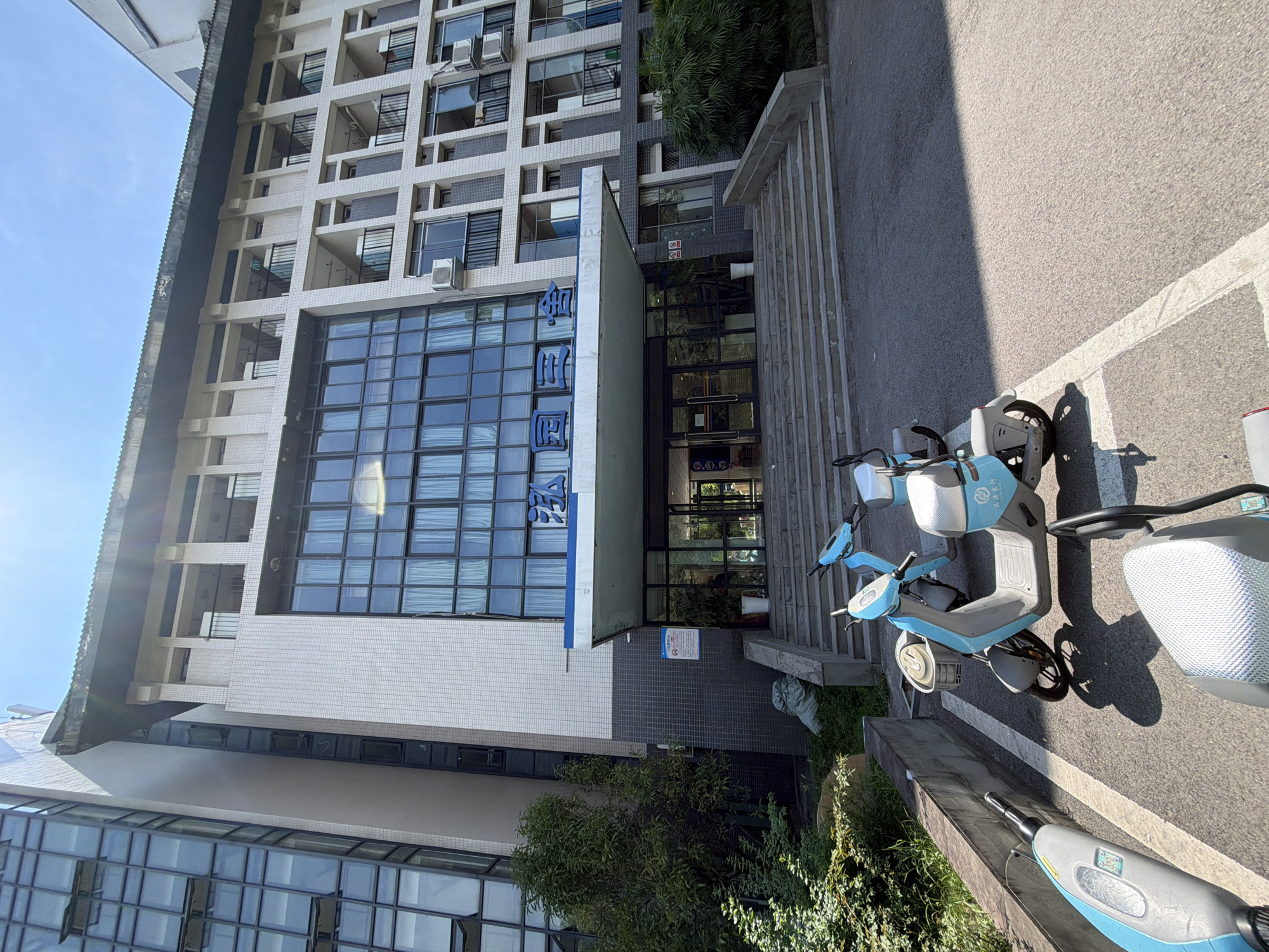 The image shows the exterior of a building, possibly an apartment complex or office building, in Jiangjin, China. The building has a modern architectural style with a facade of light-colored tiles and large glass windows. Above the entrance, blue Chinese characters are visible, spelling out "泓园三舍" which translates to "Hong Yuan San She". The entrance is flanked by potted plants and leads to a set of stairs. In front of the building, several light blue electric scooters are parked. The sky is clear and blue, suggesting a sunny day. The foreground of the image features a paved area with white parking lines.