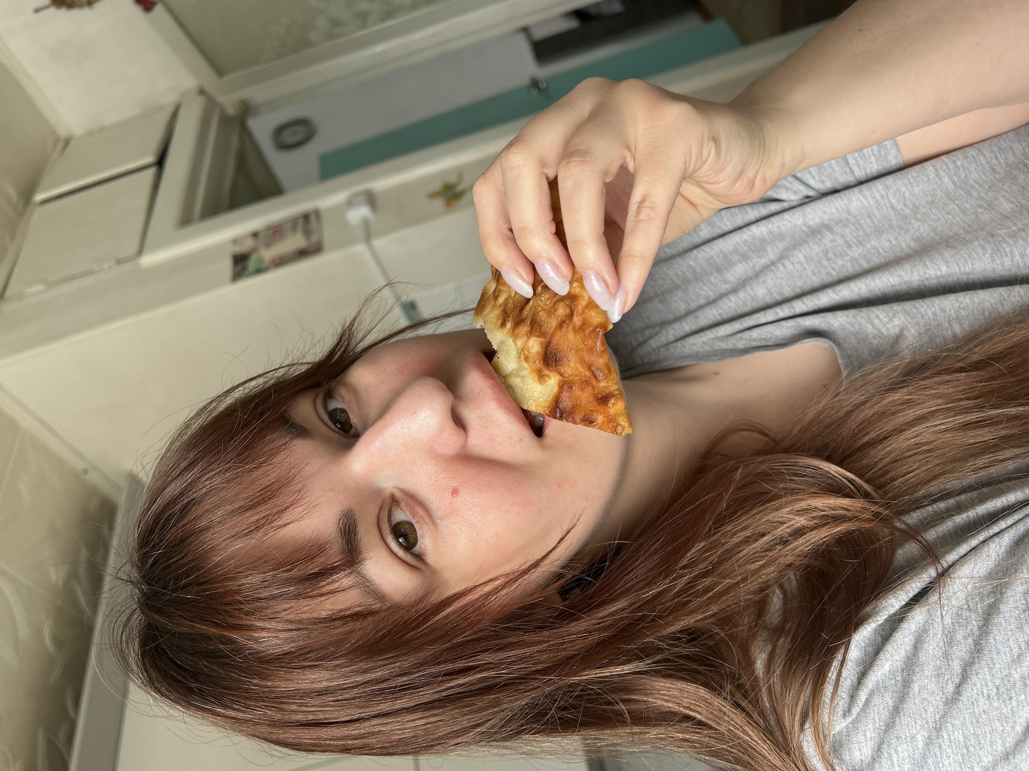 A woman with auburn hair and a gray t-shirt is holding a piece of baked food, possibly bread or pastry, to her mouth. She is looking at the camera. The background appears to be a kitchen with white cabinets.