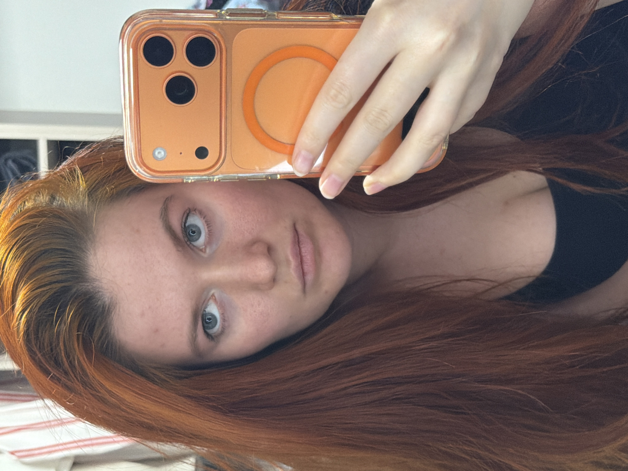 A young woman with long, reddish-brown hair is taking a selfie in an indoor setting. She is looking directly at the camera with a neutral expression. She is holding a bright orange smartphone in her right hand, which is positioned in the foreground, partially obscuring her face. The phone case is transparent with an orange magnetic ring visible on the back. She is wearing a black top. The background is slightly blurred, showing what appears to be a closet or shelving unit with some fabric items hanging on the left. The lighting suggests it is daytime, possibly from natural light coming from a window. The image does not contain any information about the location being Krasnodar, Russia.