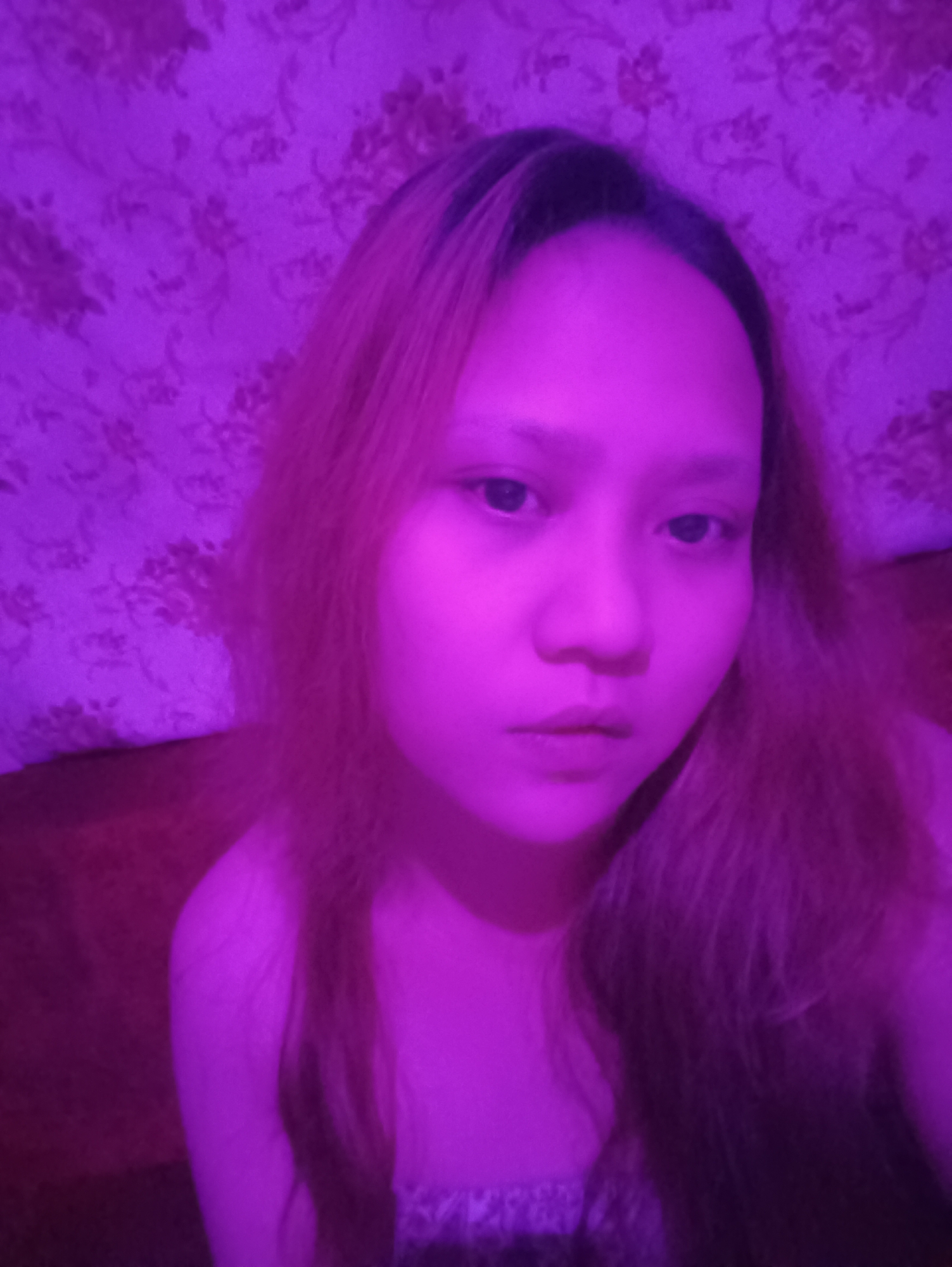 In a dimly lit room, a young woman with long, light brown hair is the central subject of the photograph. She is positioned in the foreground, her face turned slightly towards the viewer, illuminated by a strong purple light. The background features a floral patterned wallpaper, also bathed in the same purple hue. The overall scene suggests an indoor setting, potentially a bedroom or living space, in Depok, Indonesia. The woman is wearing a garment that appears to be a dress or top with a textured pattern. Her expression is neutral, and her gaze is directed forward.