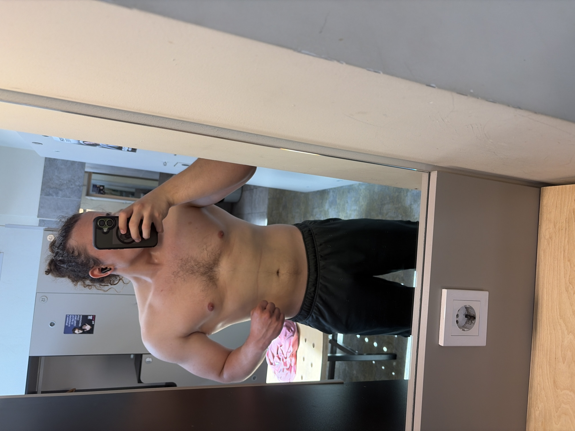 A shirtless male with curly hair stands in a locker room in Plovdiv, Bulgaria, taking a mirror selfie with a black iPhone. He is wearing black sweatpants. In the background are lockers and a table with a pink object. There is an electrical outlet on the wall to the right of the mirror.
